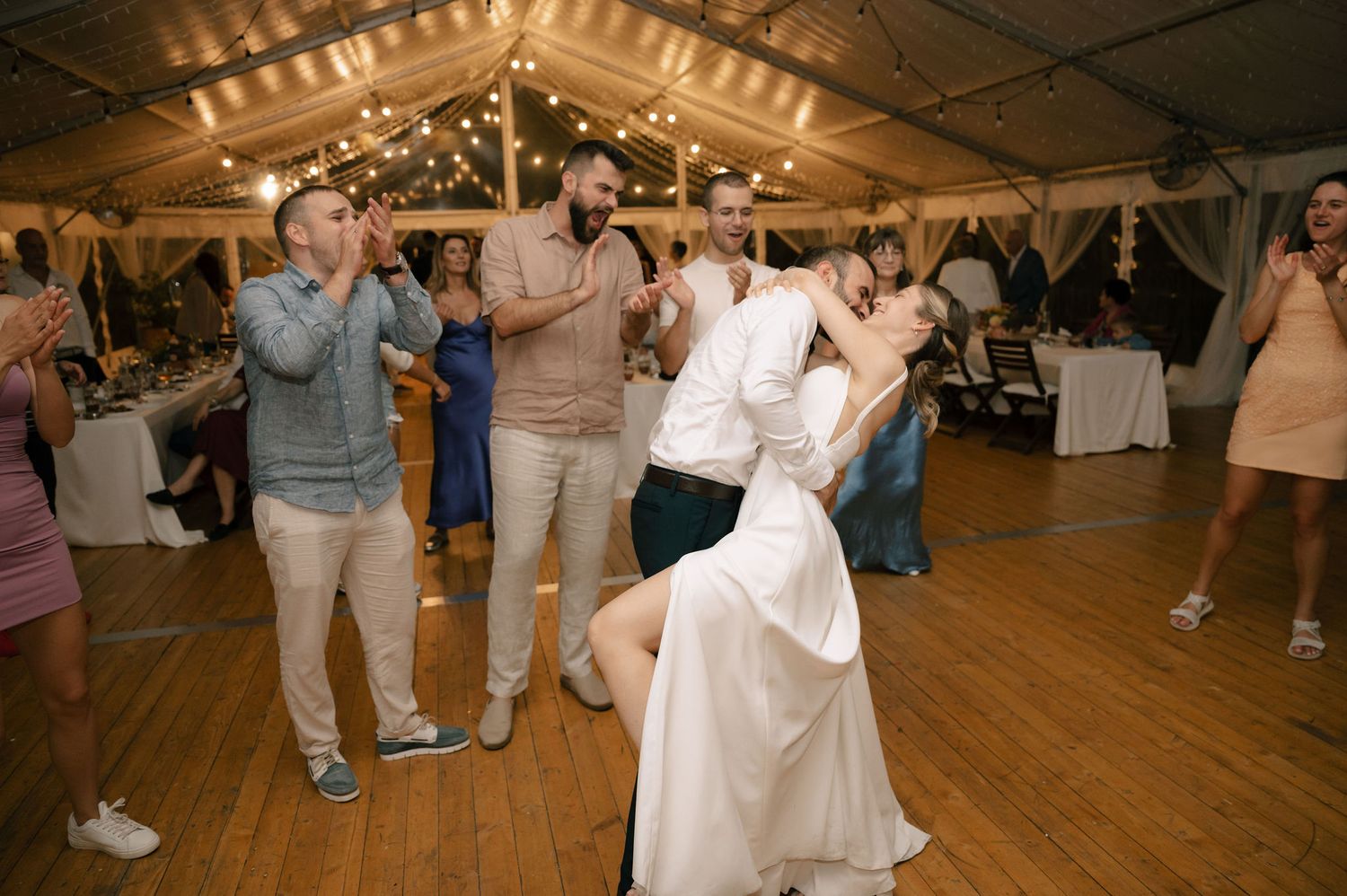 People dancing and celebrating at a wedding reception on a wooden dance floor under tent lighting.