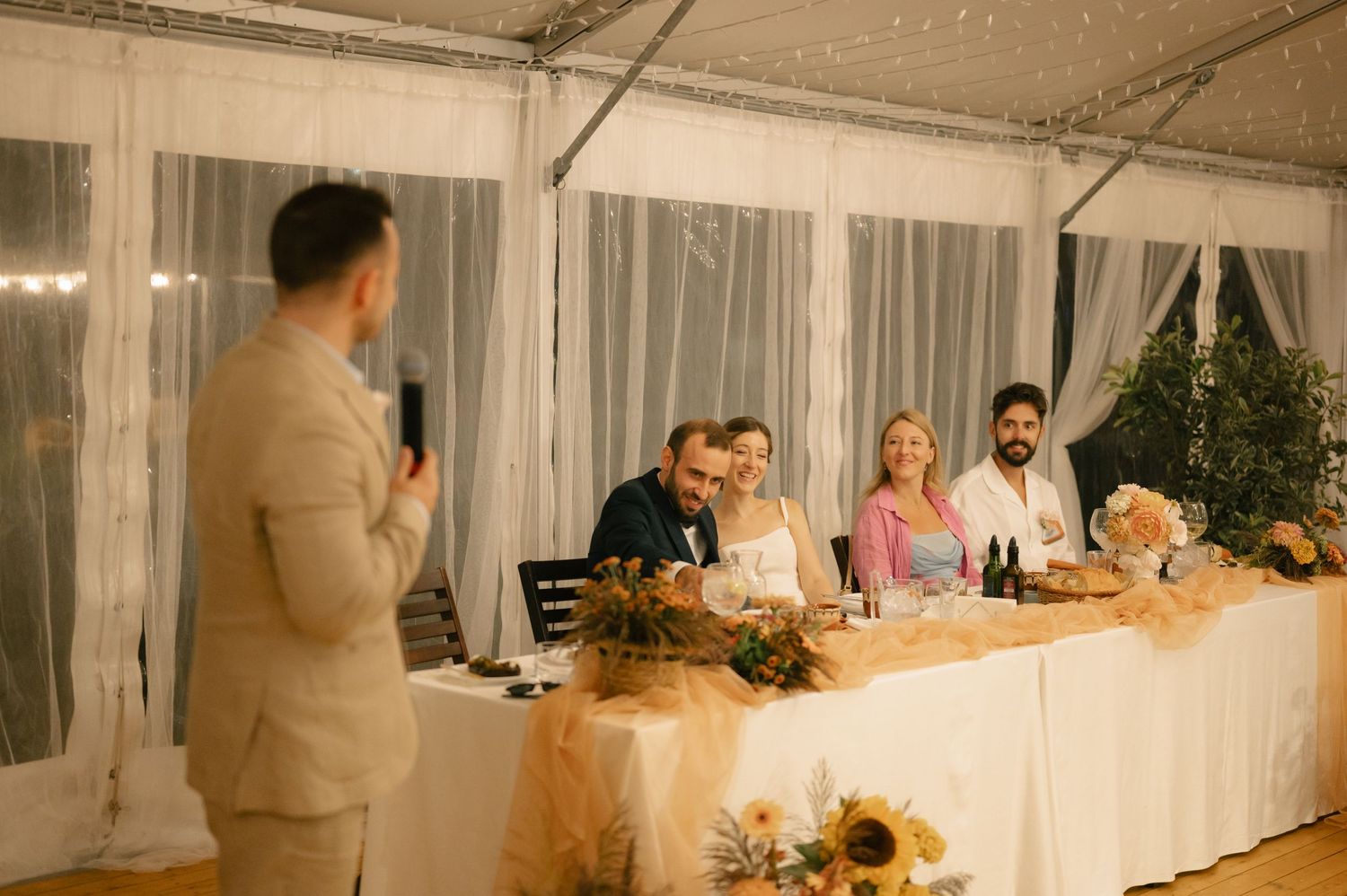 Wedding reception venue decorated with string lights and white linens features guests mingling at tables.