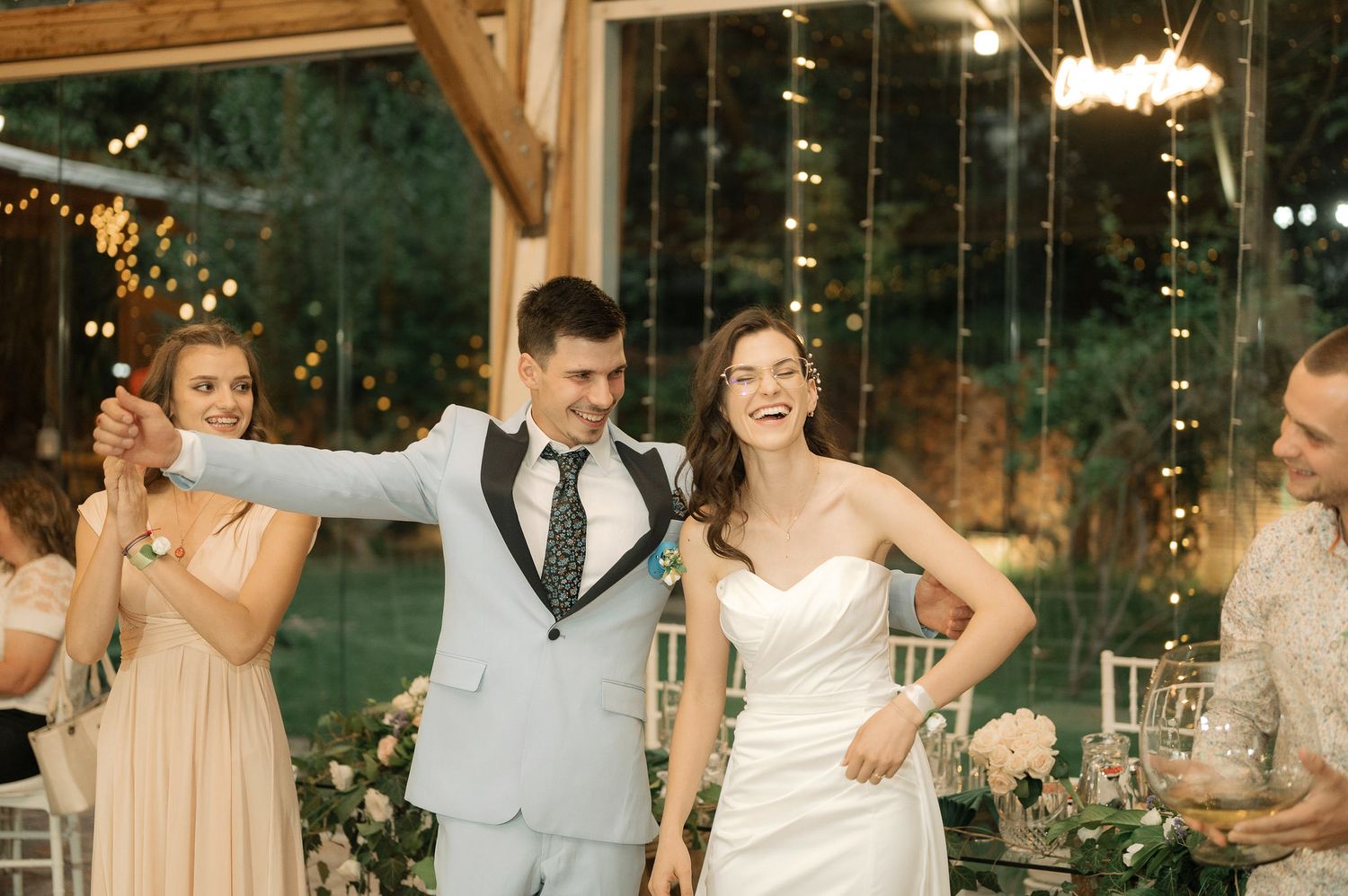 Wedding party celebrates joyfully during an outdoor evening reception with string lights and greenery.