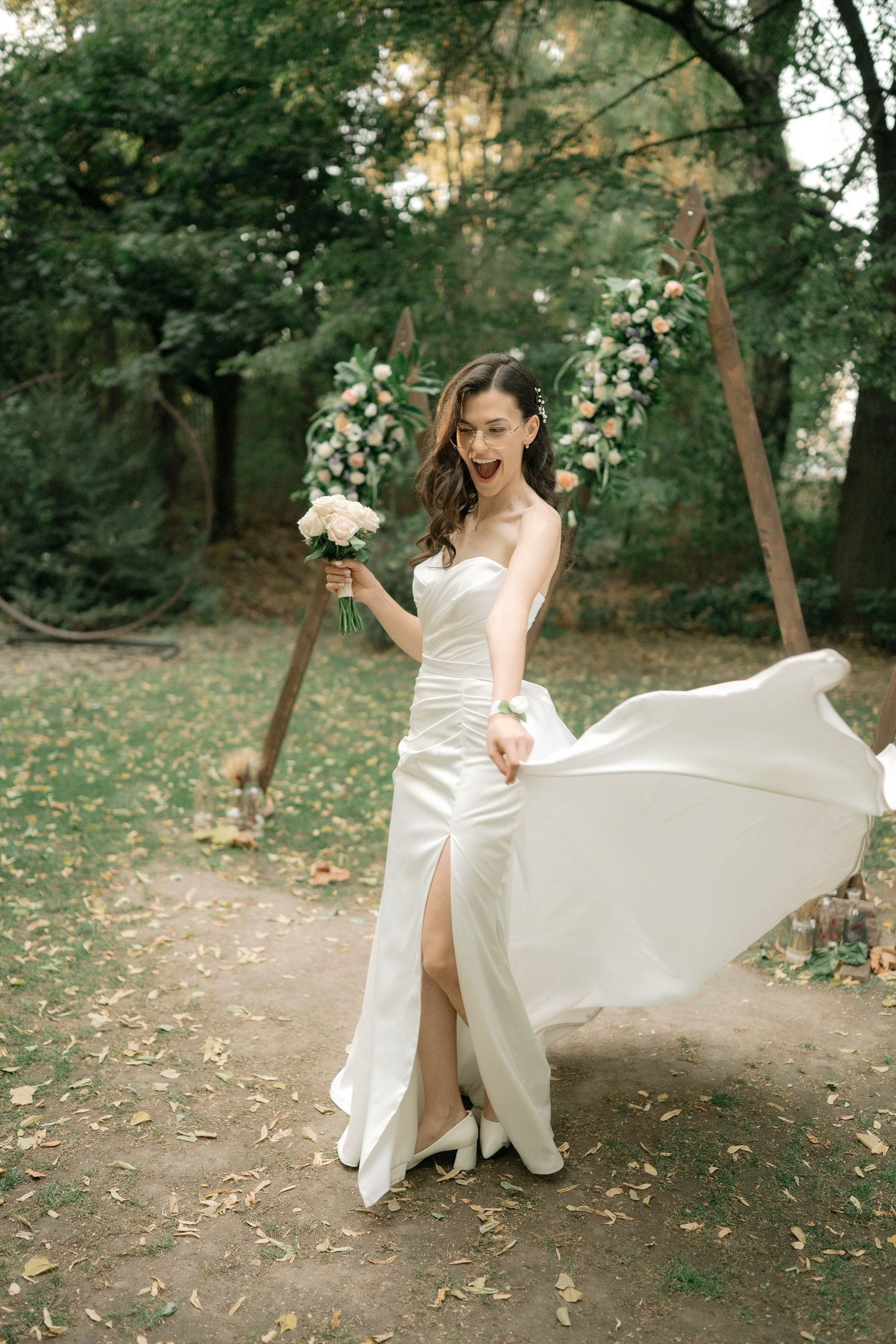 A bride in a flowing white wedding dress twirls playfully in a wooded outdoor setting during a photo session.