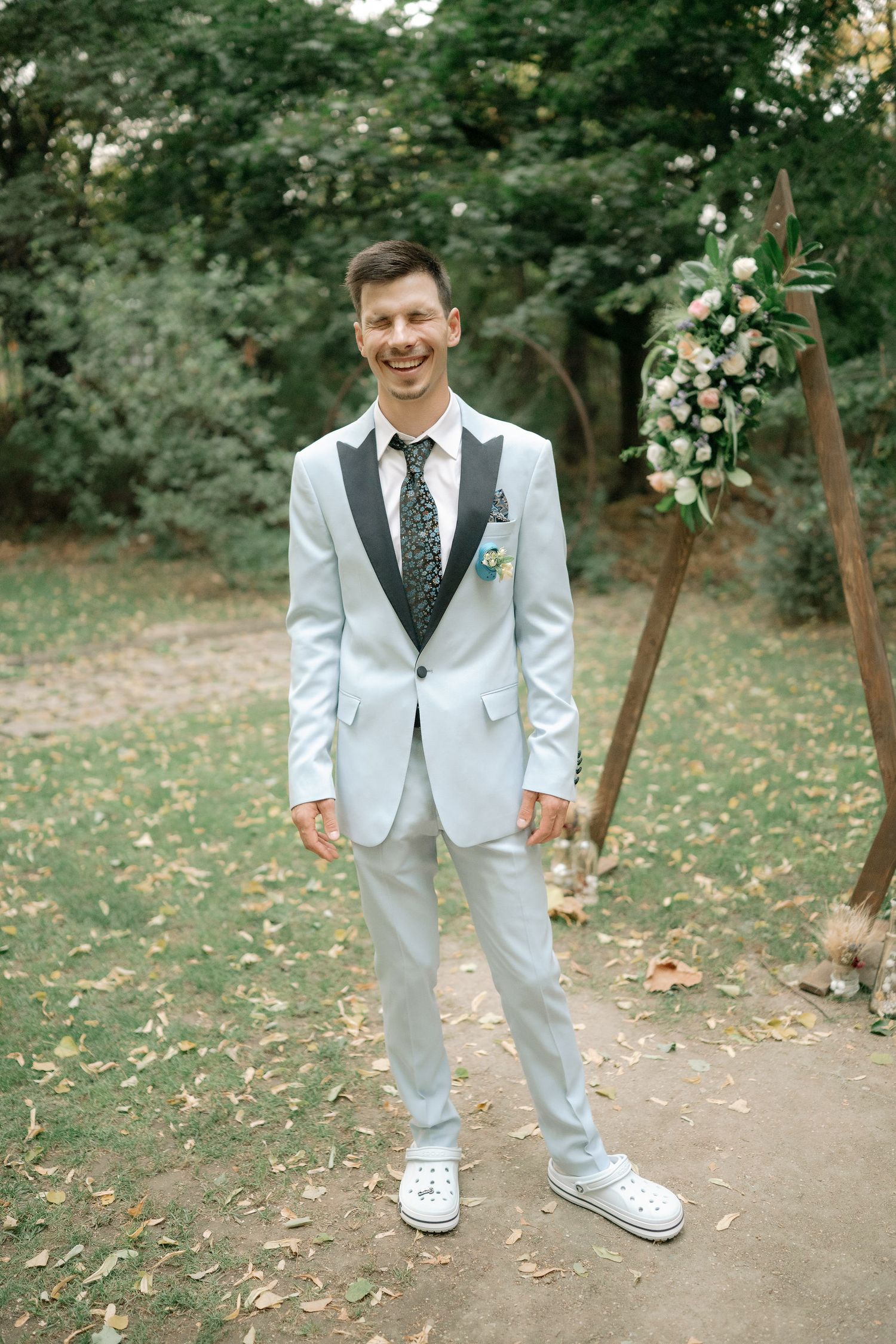 A person wearing a light gray suit with black lapels and white sneakers stands on a path surrounded by green trees.