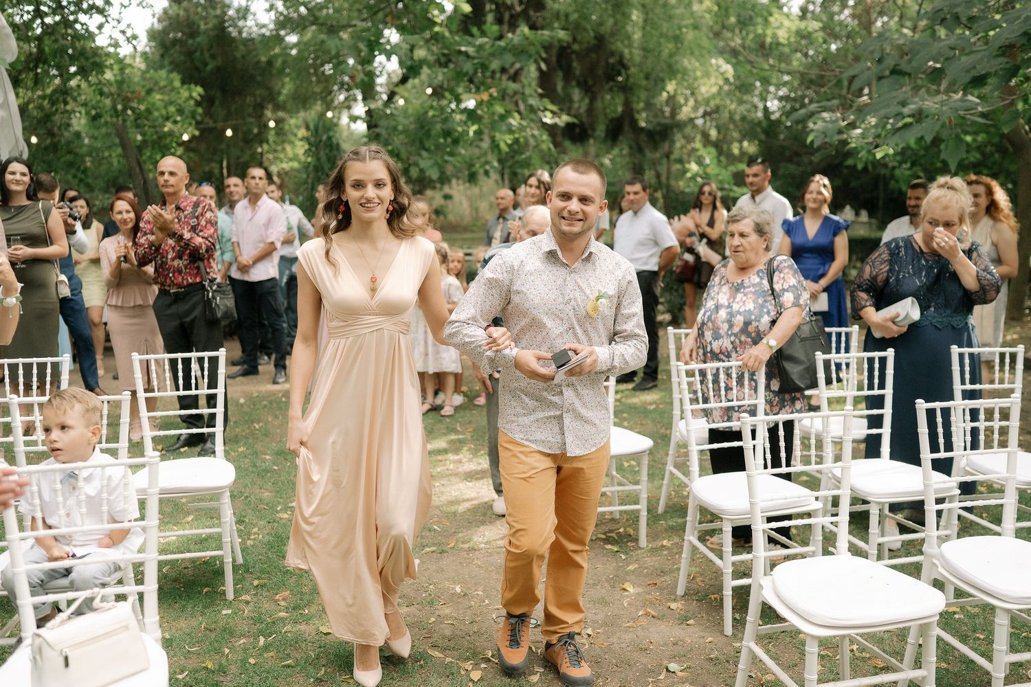 White chairs arranged outdoors for a garden wedding ceremony with guests arriving in pastel attire.