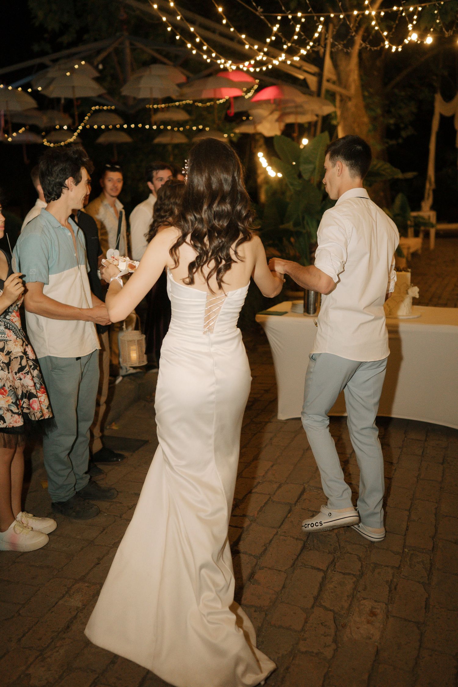 Wedding guests dance together on a brick patio under twinkling string lights during an evening reception celebration.