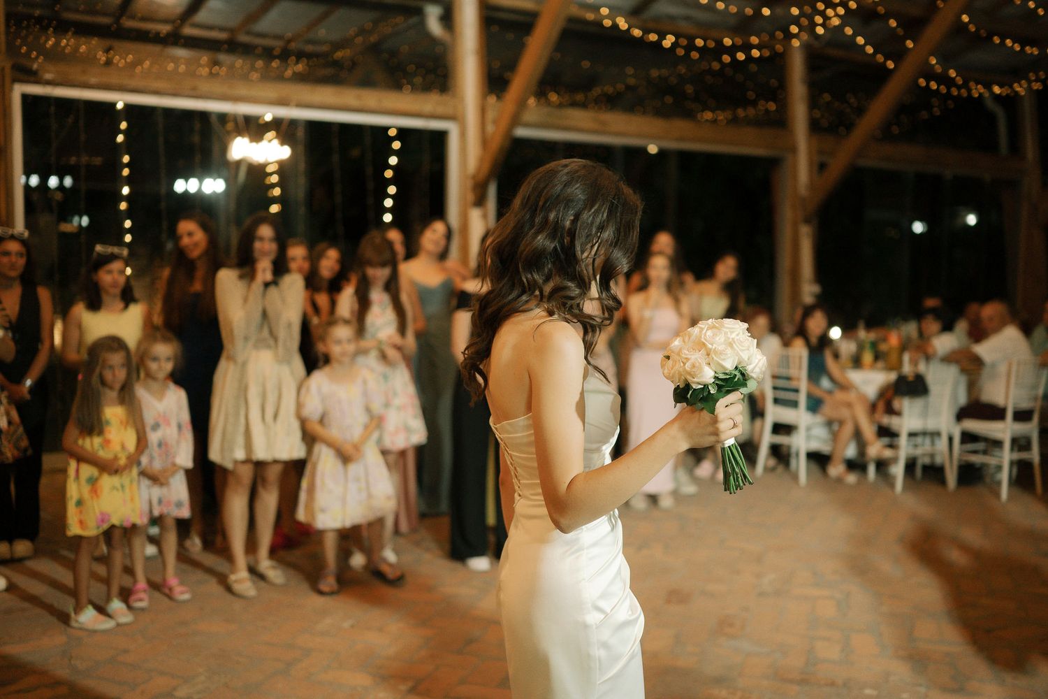 A bride in a white dress stands in a rustic venue with string lights while guests gather in the background.