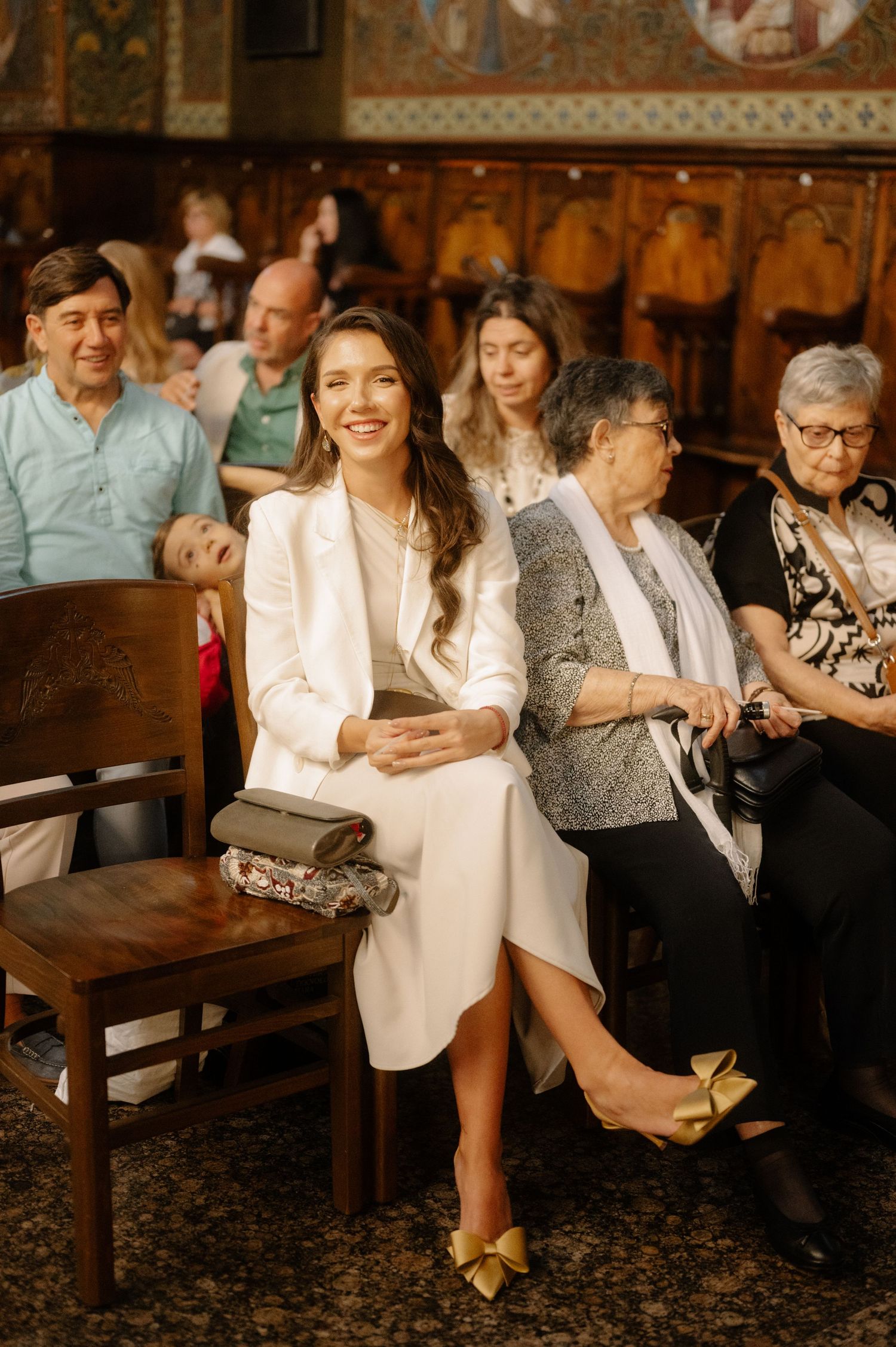 People seated in wooden pews inside an ornate church or chapel with decorative walls and vintage furnishings.
