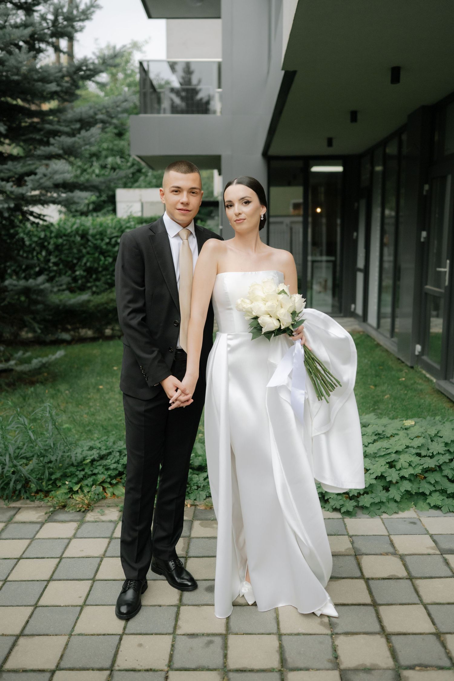 A couple poses together outdoors on their wedding day, with the bride in white holding a bouquet and groom in dark suit.
