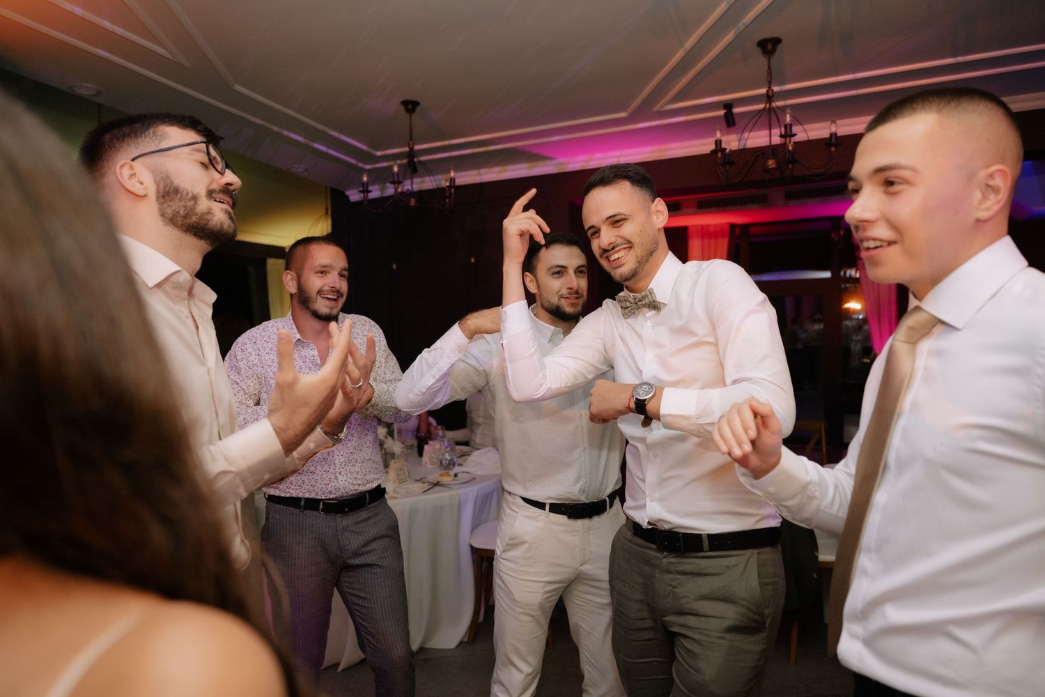 Wedding guests in white shirts and gray pants dancing together at a reception with pink lighting.