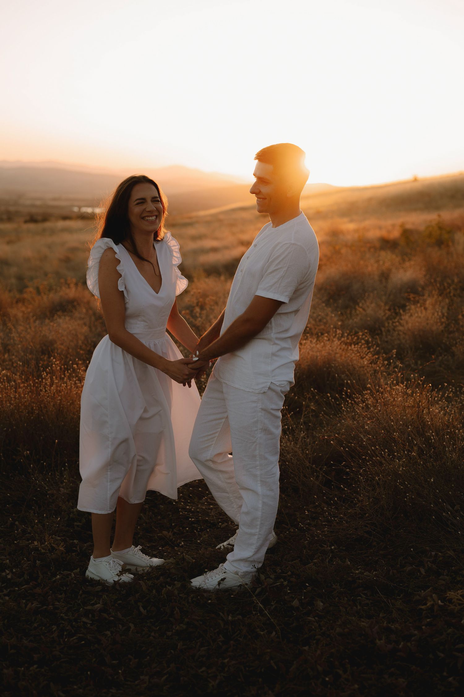 A couple wearing white outfits shares an intimate moment at sunset in a golden field.