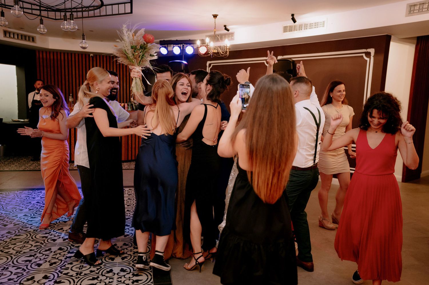 People dancing and celebrating at an indoor event with decorative lighting and a patterned carpet floor.