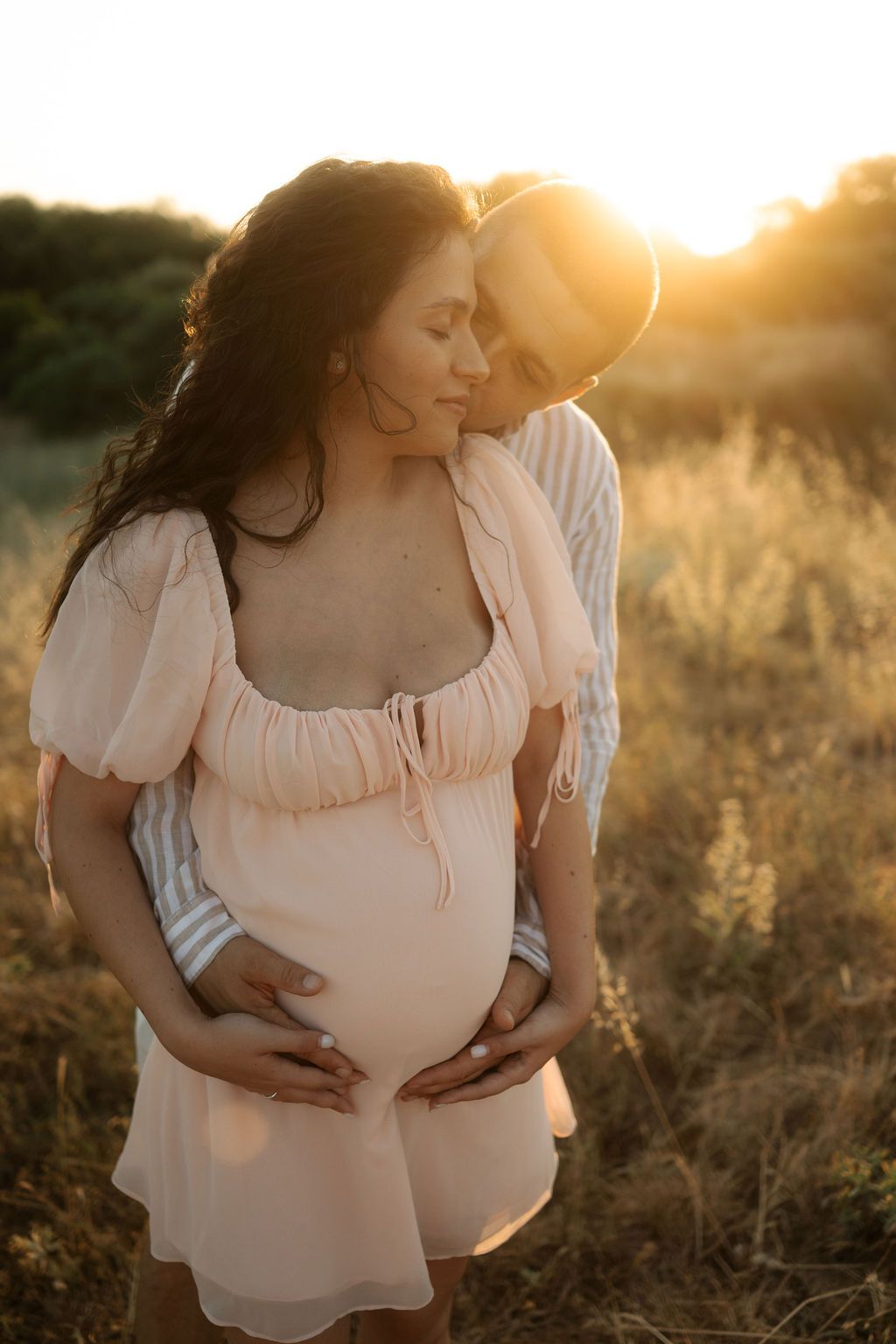 Couple embraces in golden sunset light while standing in tall grass field wearing white and cream colored clothing.