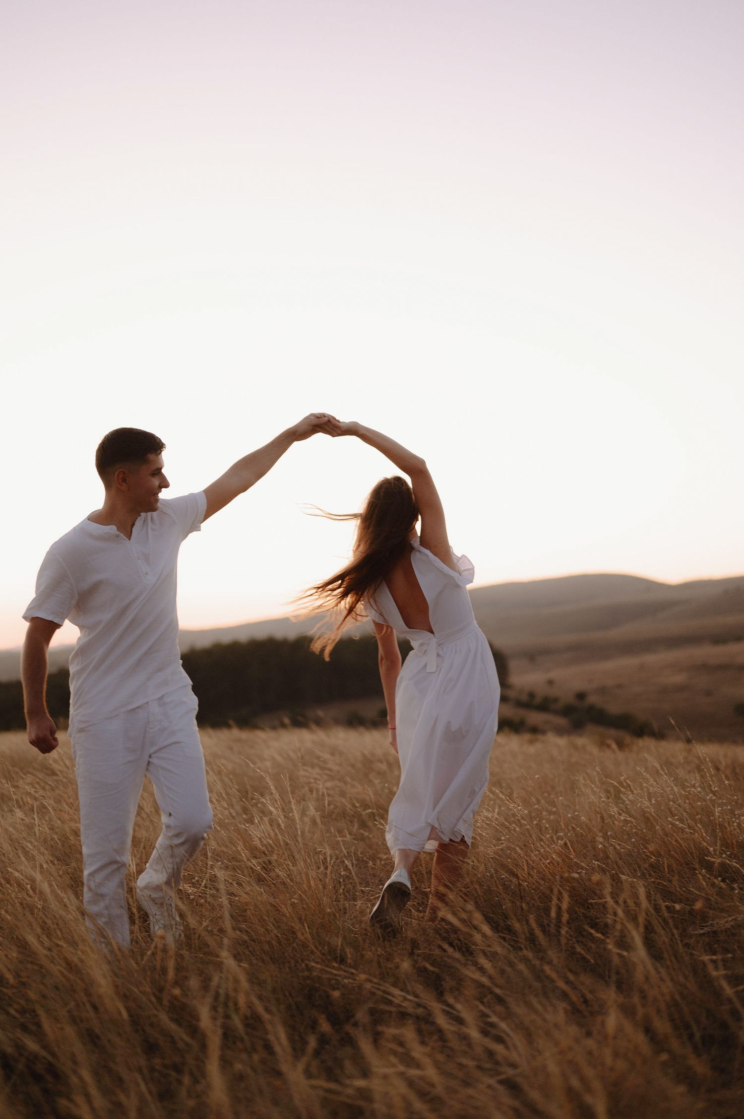 A couple in white outfits dancing together in a golden wheat field at sunset.