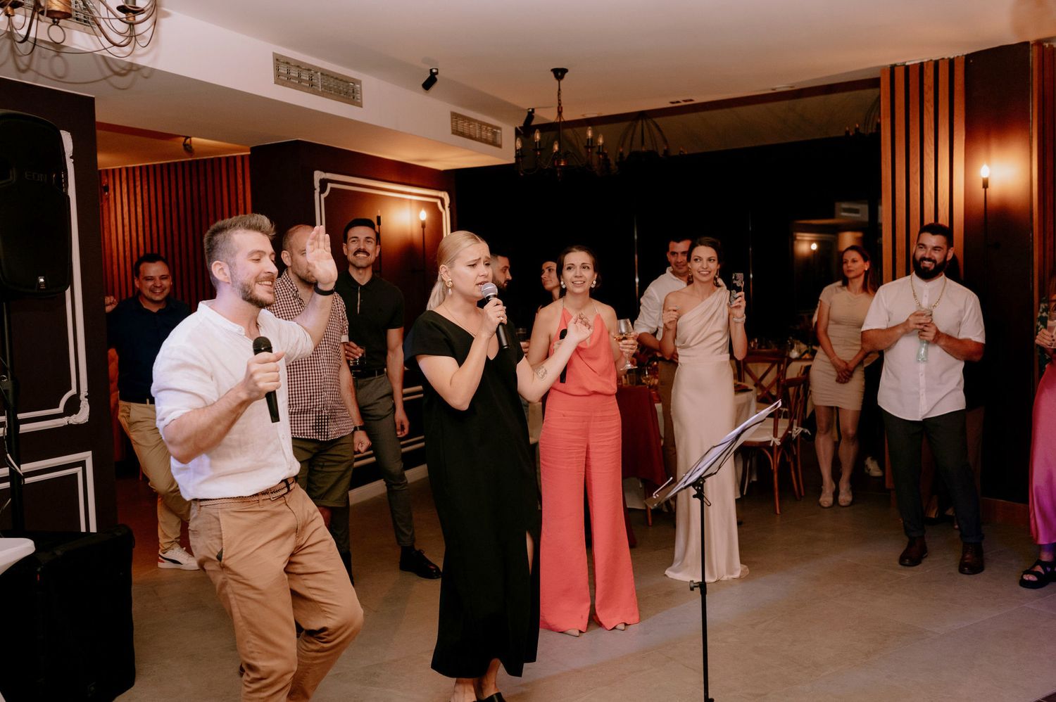 A group of party guests dance and celebrate together in a warmly lit indoor venue during an evening event.
