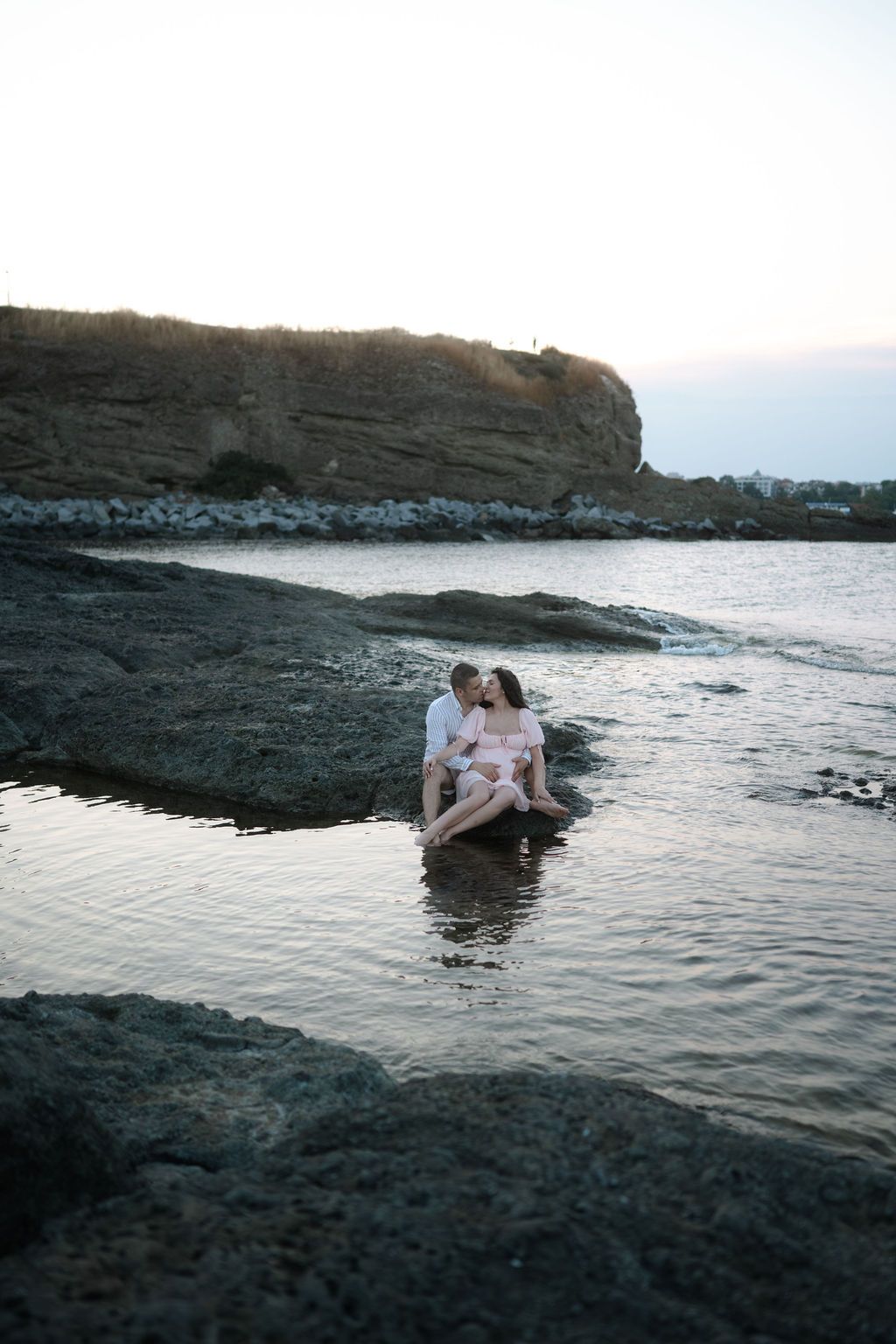A couple embraces while sitting on dark volcanic rocks at the ocean's edge during sunset.