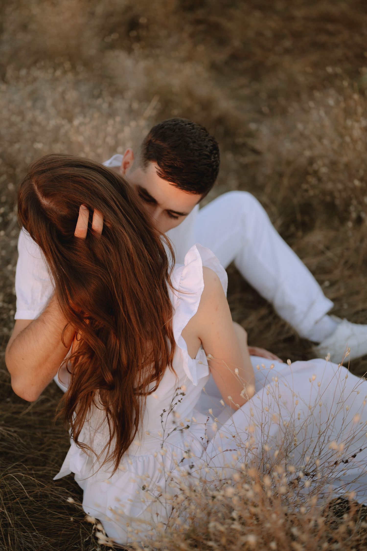 Romantic couple in white clothing embraces while sitting in a field of dried grass during golden hour.