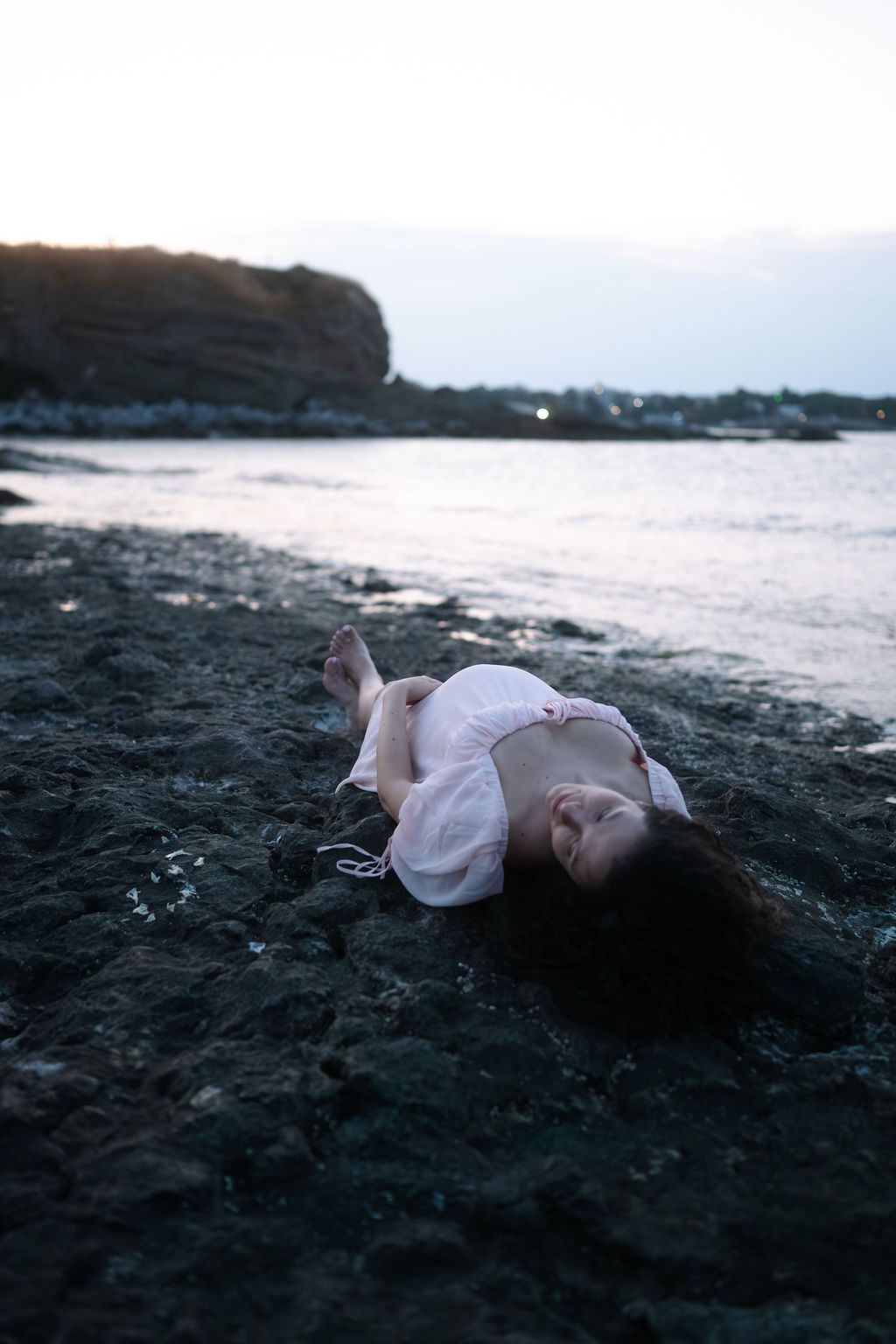A person in white clothing lies peacefully on dark volcanic sand at the ocean's edge during sunset.