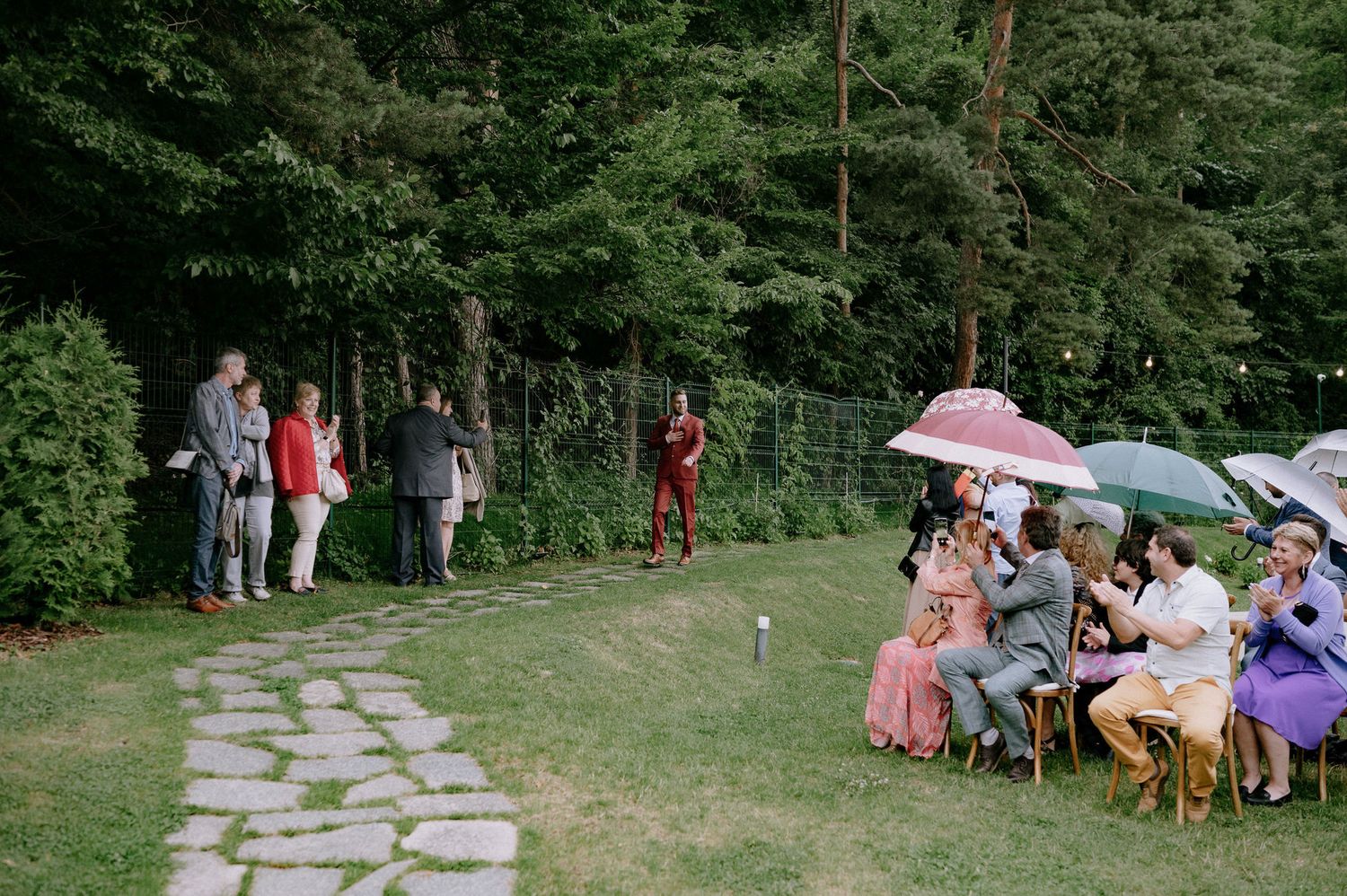 Wedding guests gather outdoors on a stone path and lawn area with umbrellas during a garden ceremony.