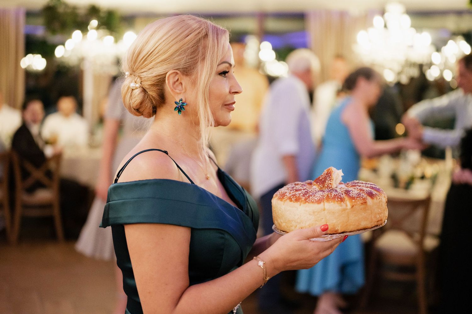 A person in an elegant navy blue dress holds a decorated birthday cake at an upscale event with chandeliers in the background.