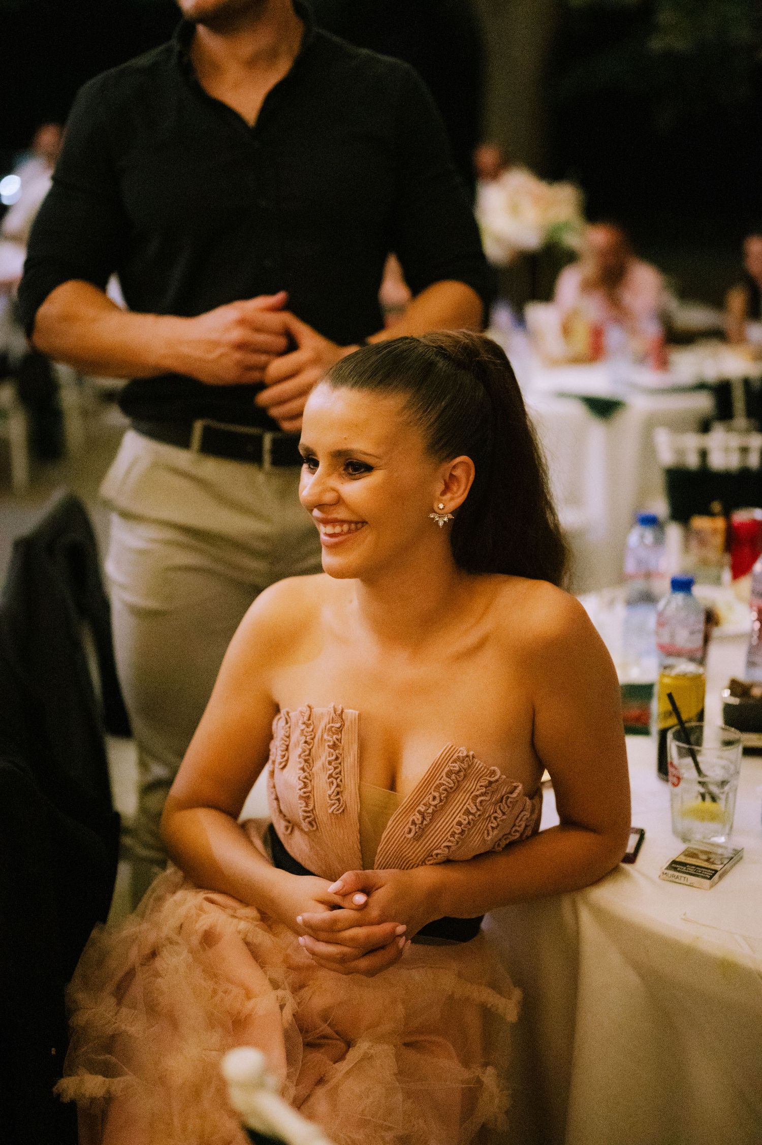 Woman in strapless blush gown smiles while seated at elegant reception table during evening event.