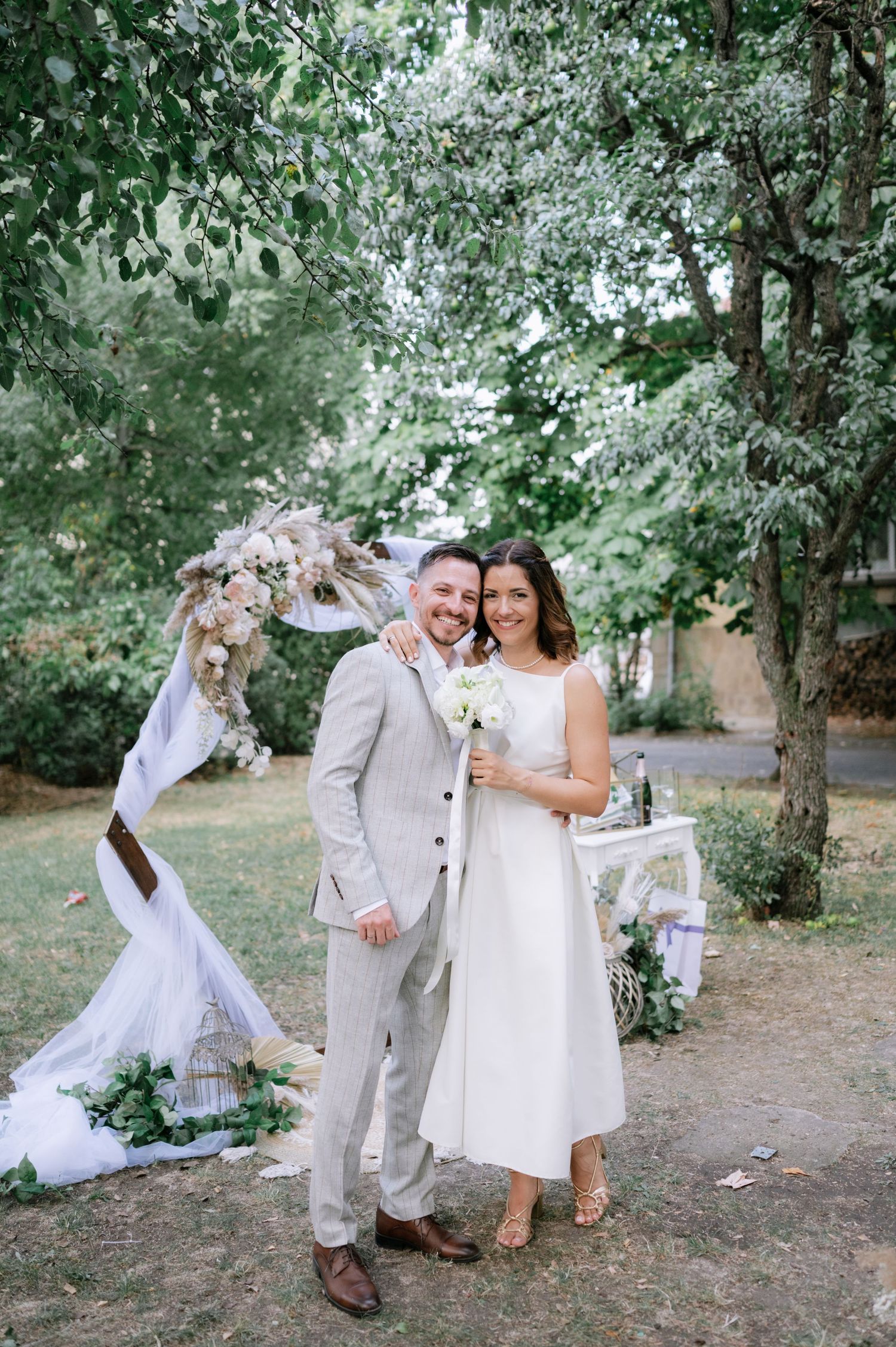 A couple in white outfits shares joyful moments during their outdoor garden wedding photoshoot.