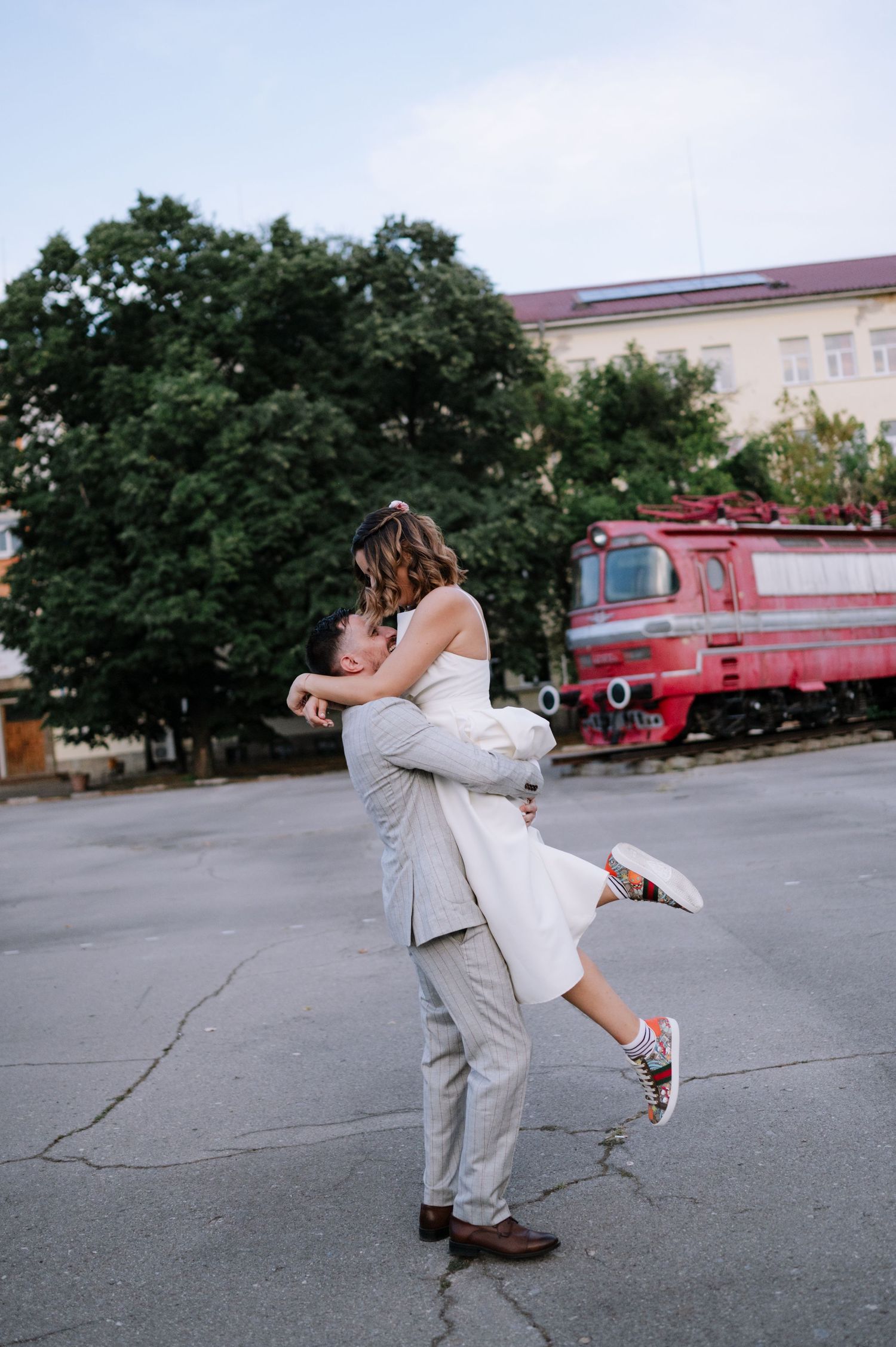 A romantic couple embraces in front of a red train at a train station, with the person in white being lifted off the ground.