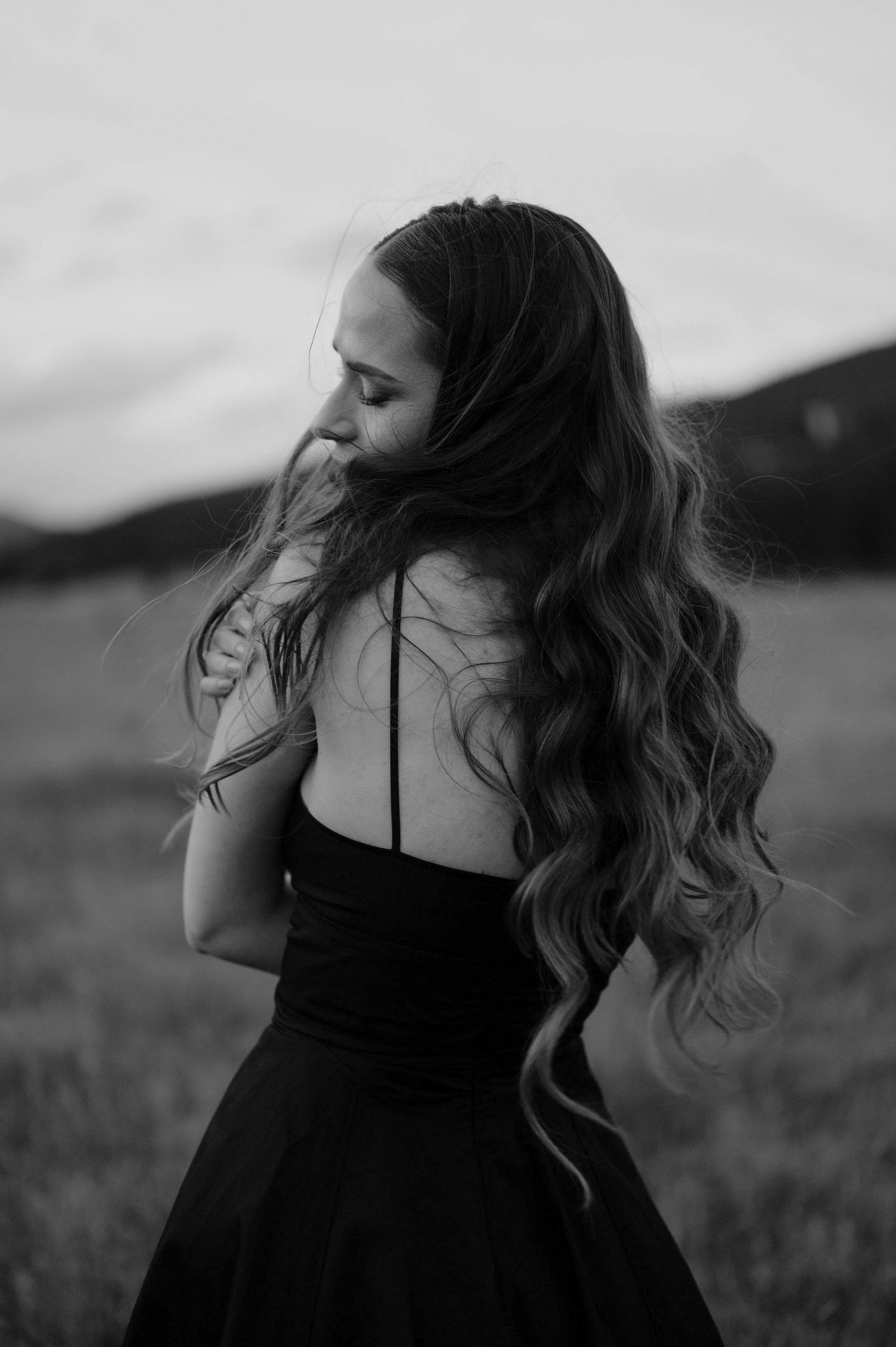 A woman in a black dress stands in a field at dusk with her hair flowing in the wind in a black and white photo.