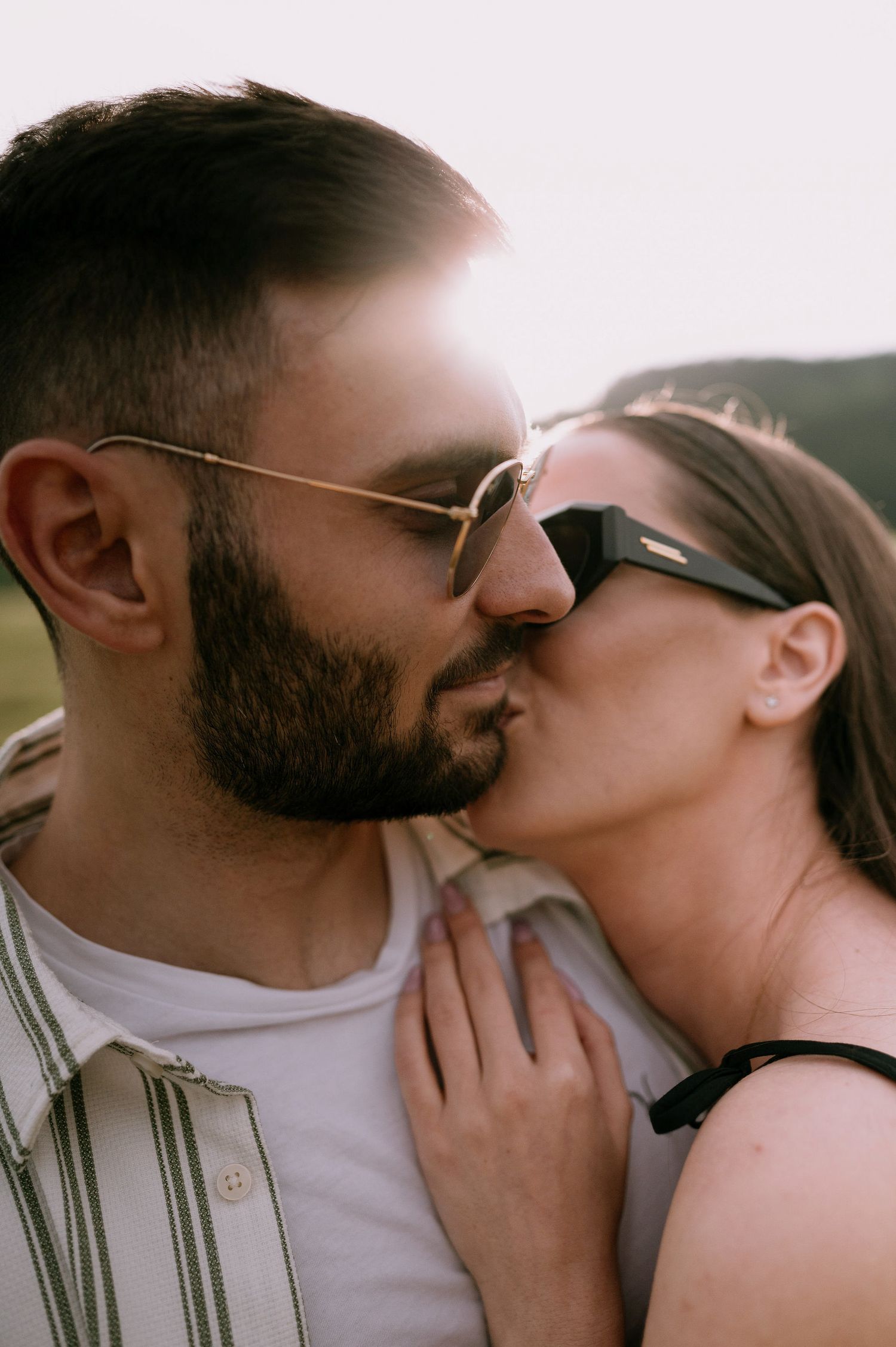 A romantic couple sharing an intimate moment outdoors, wearing sunglasses and embracing in soft sunlight.