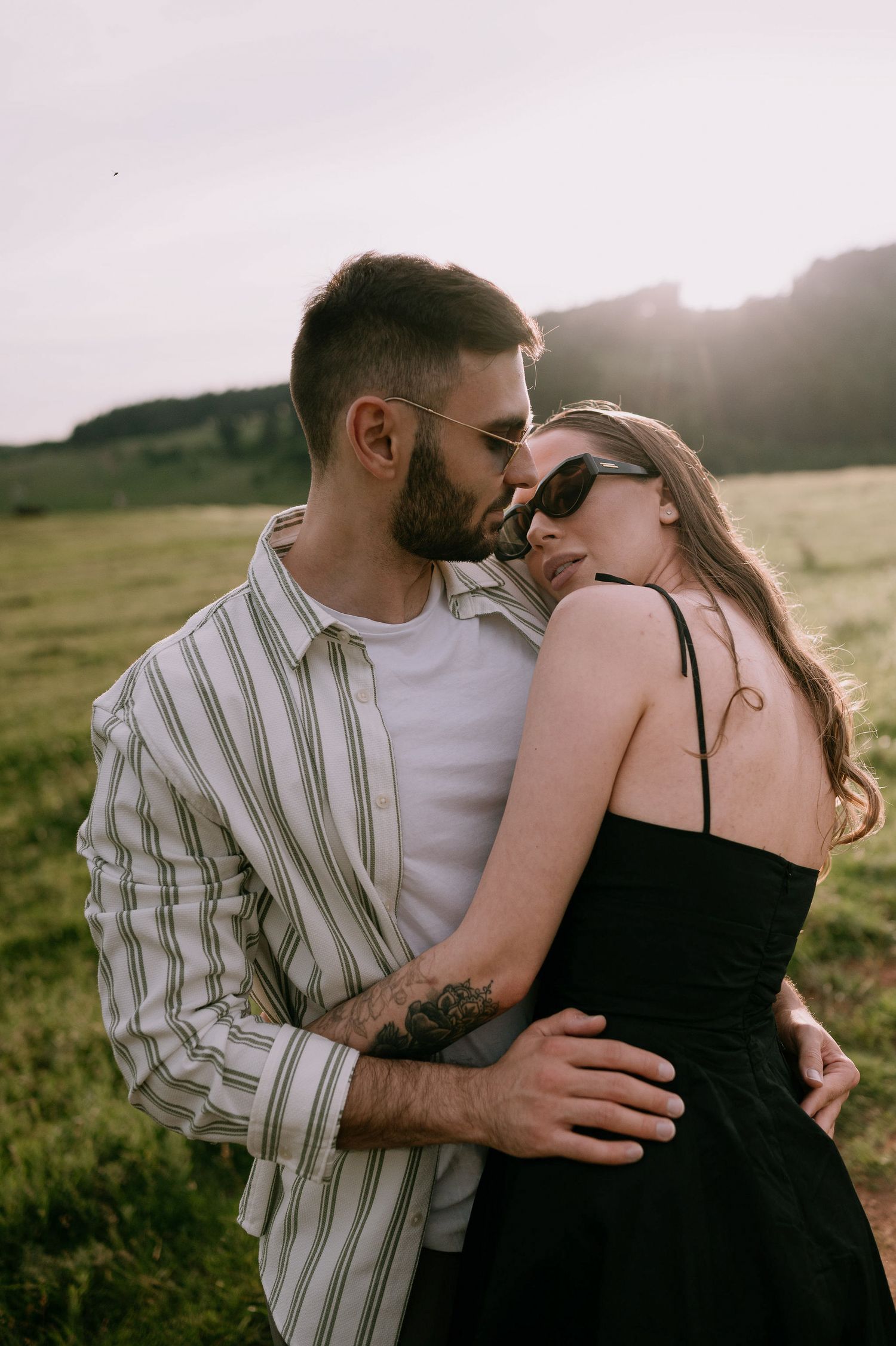 A romantic couple embraces in a grassy field at sunset wearing a striped shirt and black dress.