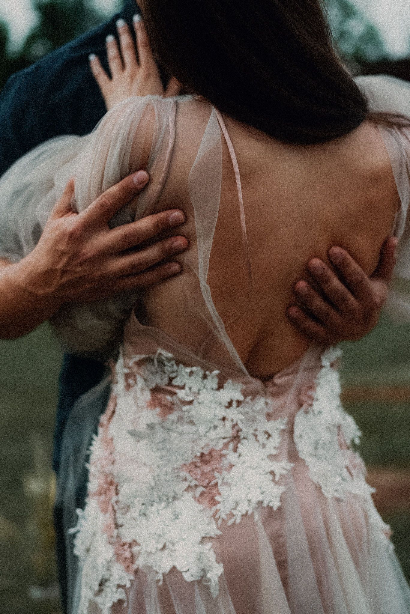 Intimate wedding photo showing the delicate lace back detail of a sheer white bridal gown with hands embracing.