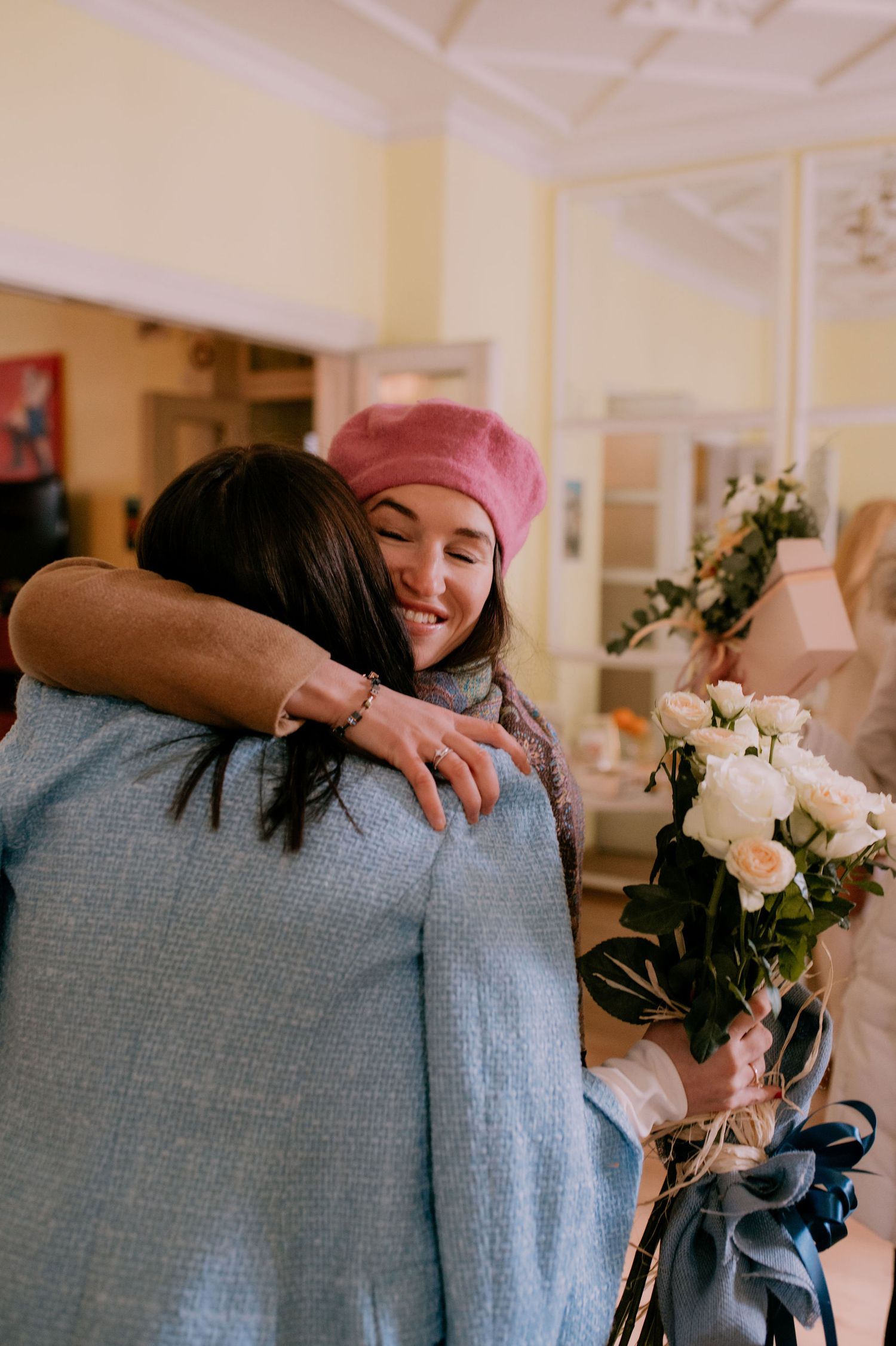 Two people share an emotional embrace in a hallway while one holds a bouquet of pink and white flowers.