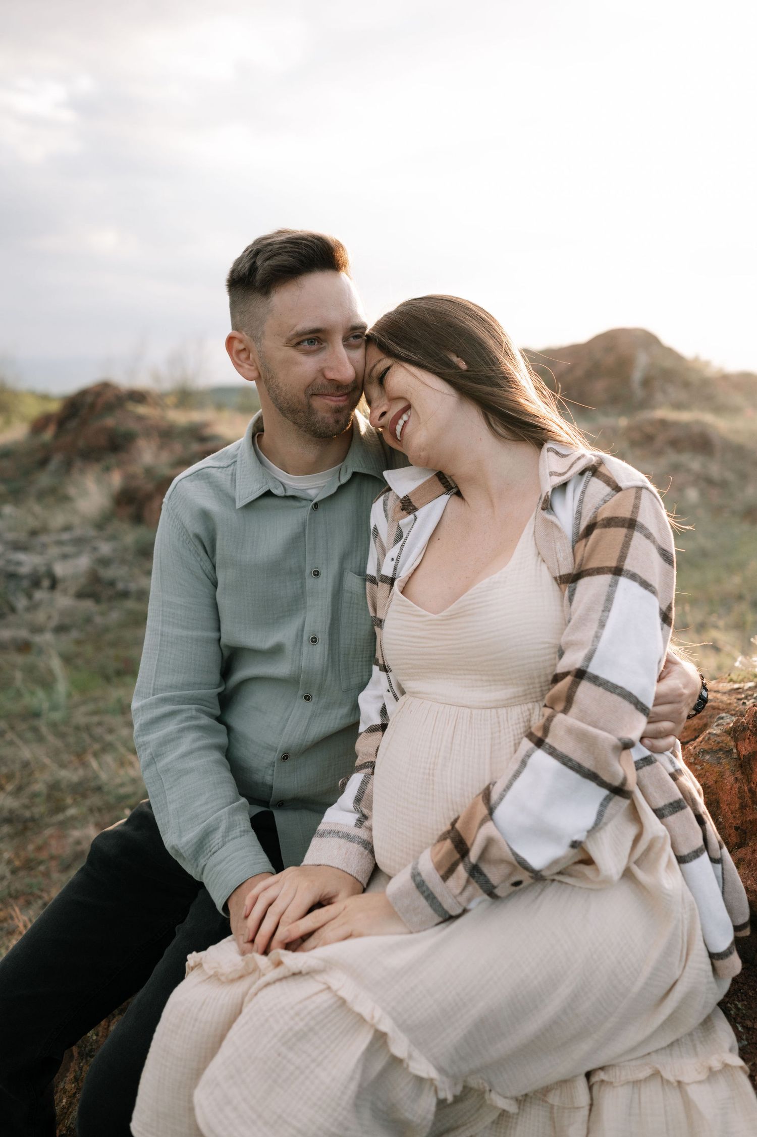 A couple shares an intimate moment outdoors at sunset while sitting together on natural terrain.