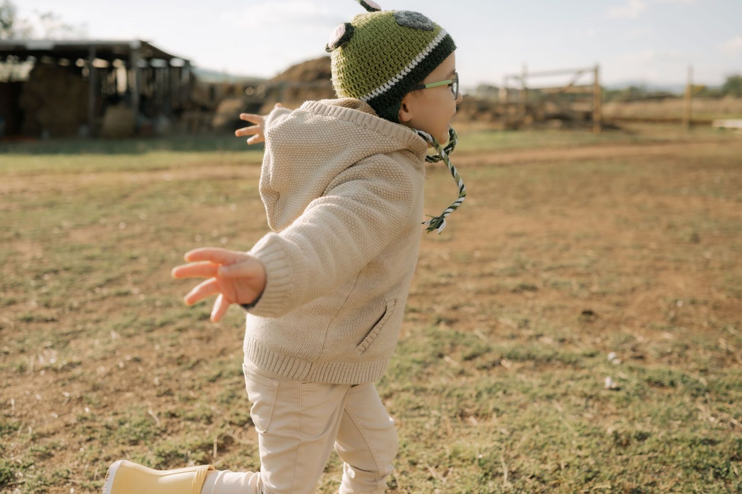 A child wearing a beige outfit and knit hat walks across a dry grassy field on an autumn day.