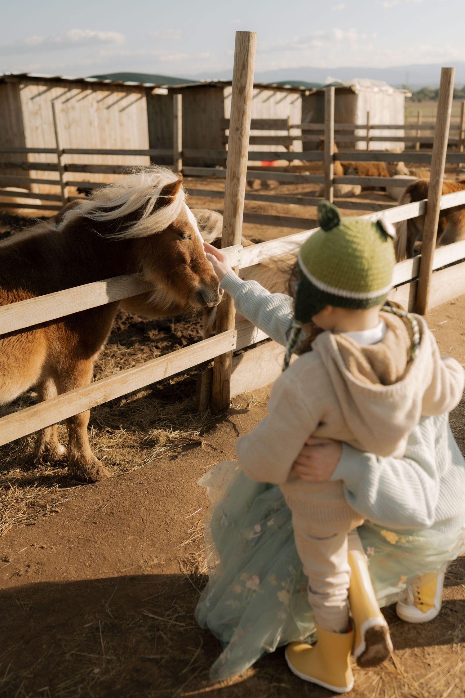 Two children in white outfits and hats sit on a wooden fence at a ranch on a sunny day.