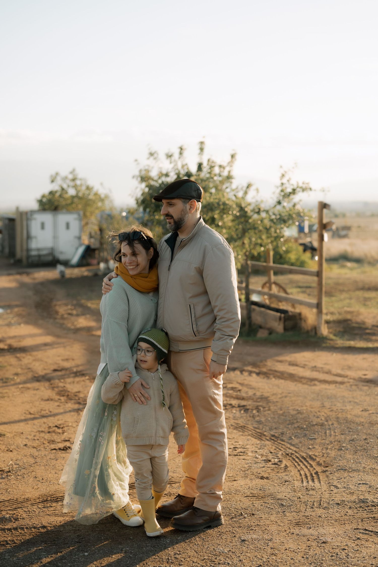Family in warm autumn clothing stands together on a dirt path at sunset in a rural setting.