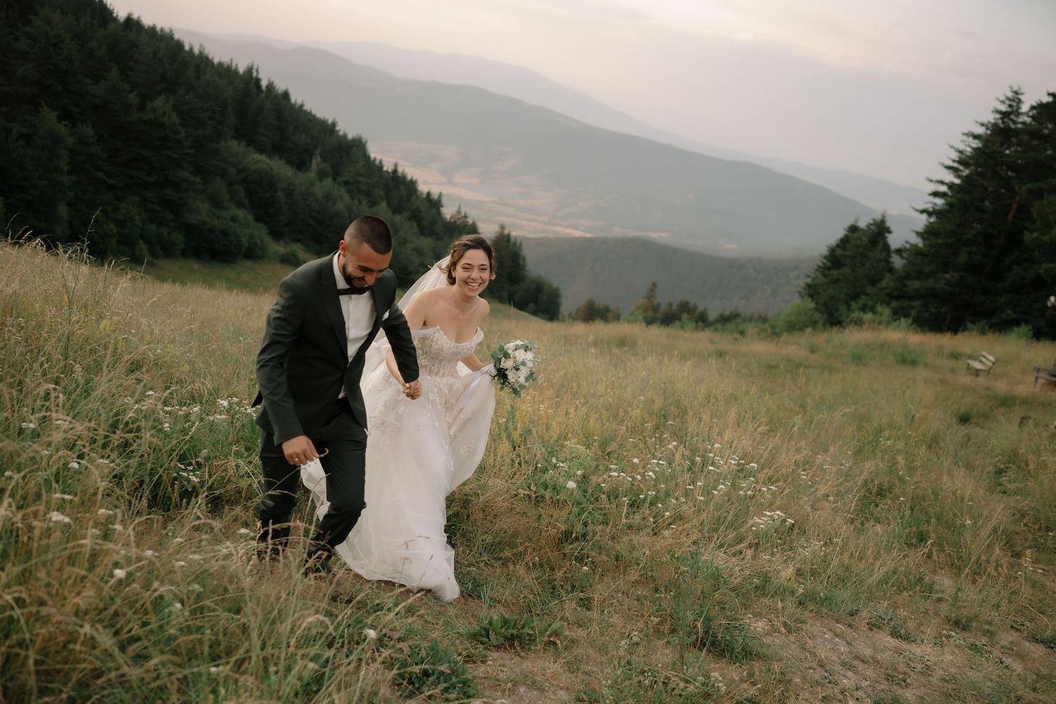 A couple shares romantic moments in a scenic mountain meadow with wildflowers and evergreen trees in the background.