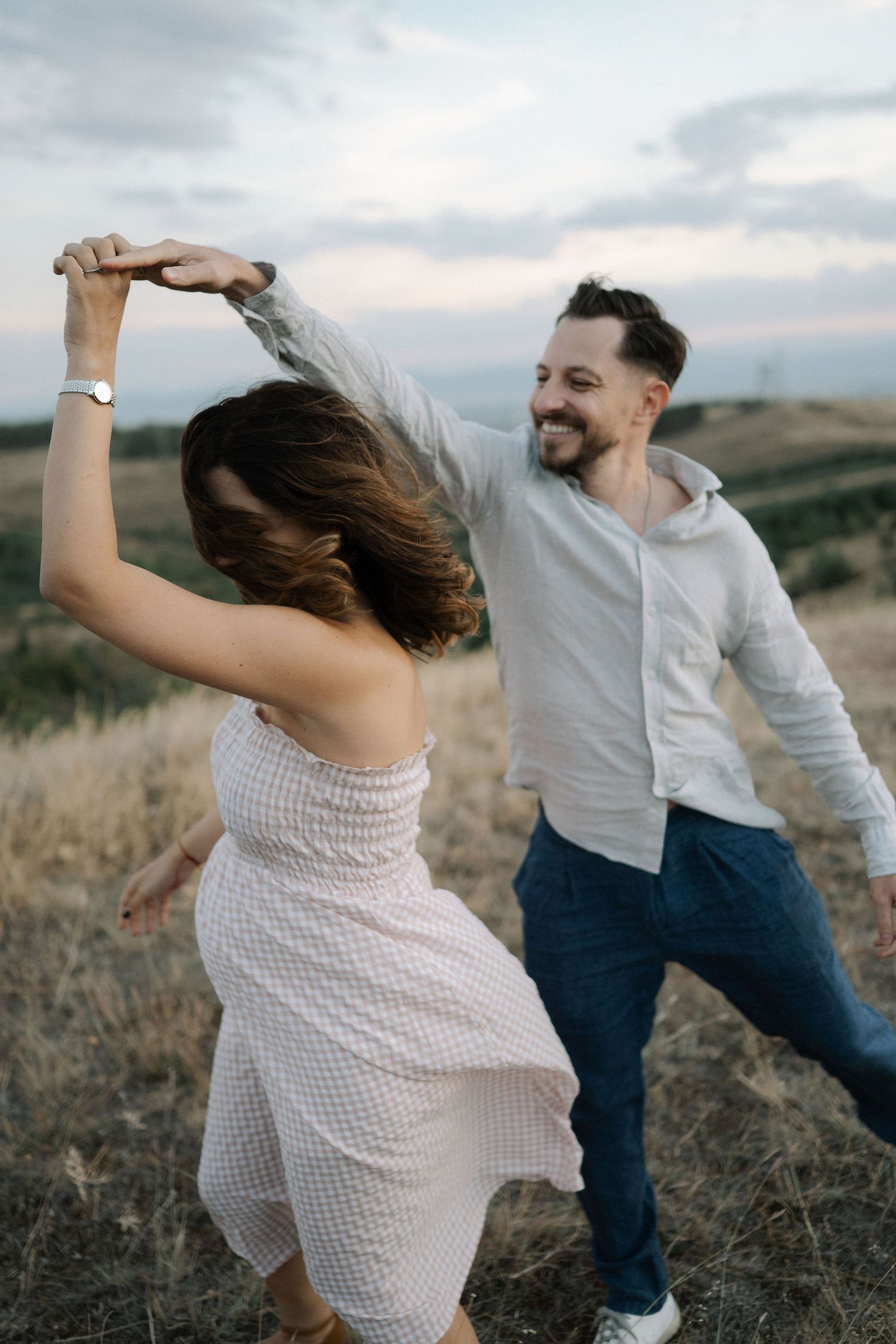 A couple dances together joyfully in a field at sunset, wearing casual white and blue outfits.