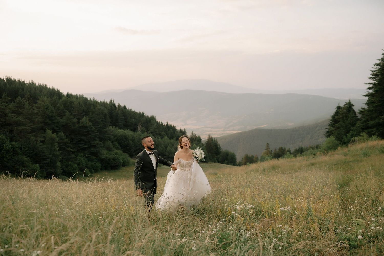 A couple walks through a scenic mountain meadow at sunset with rolling hills and forests in the background.