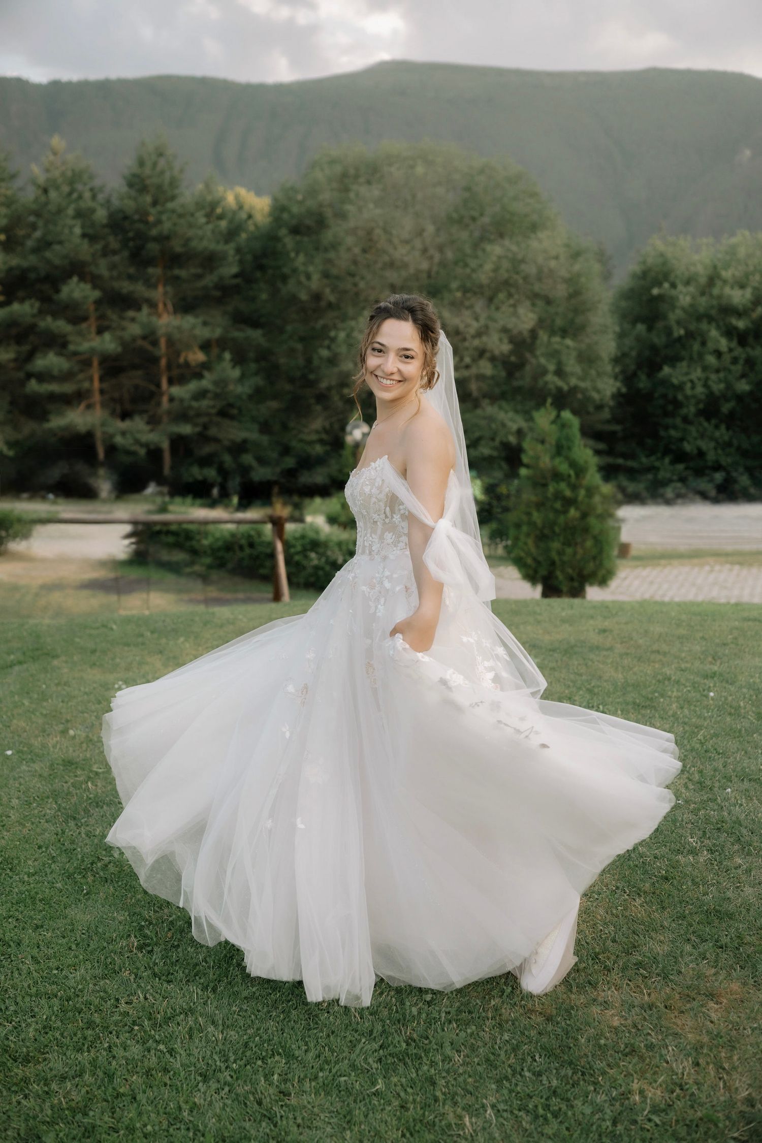 A flowing white wedding dress twirls gracefully on a grassy hillside with mountains in the background.