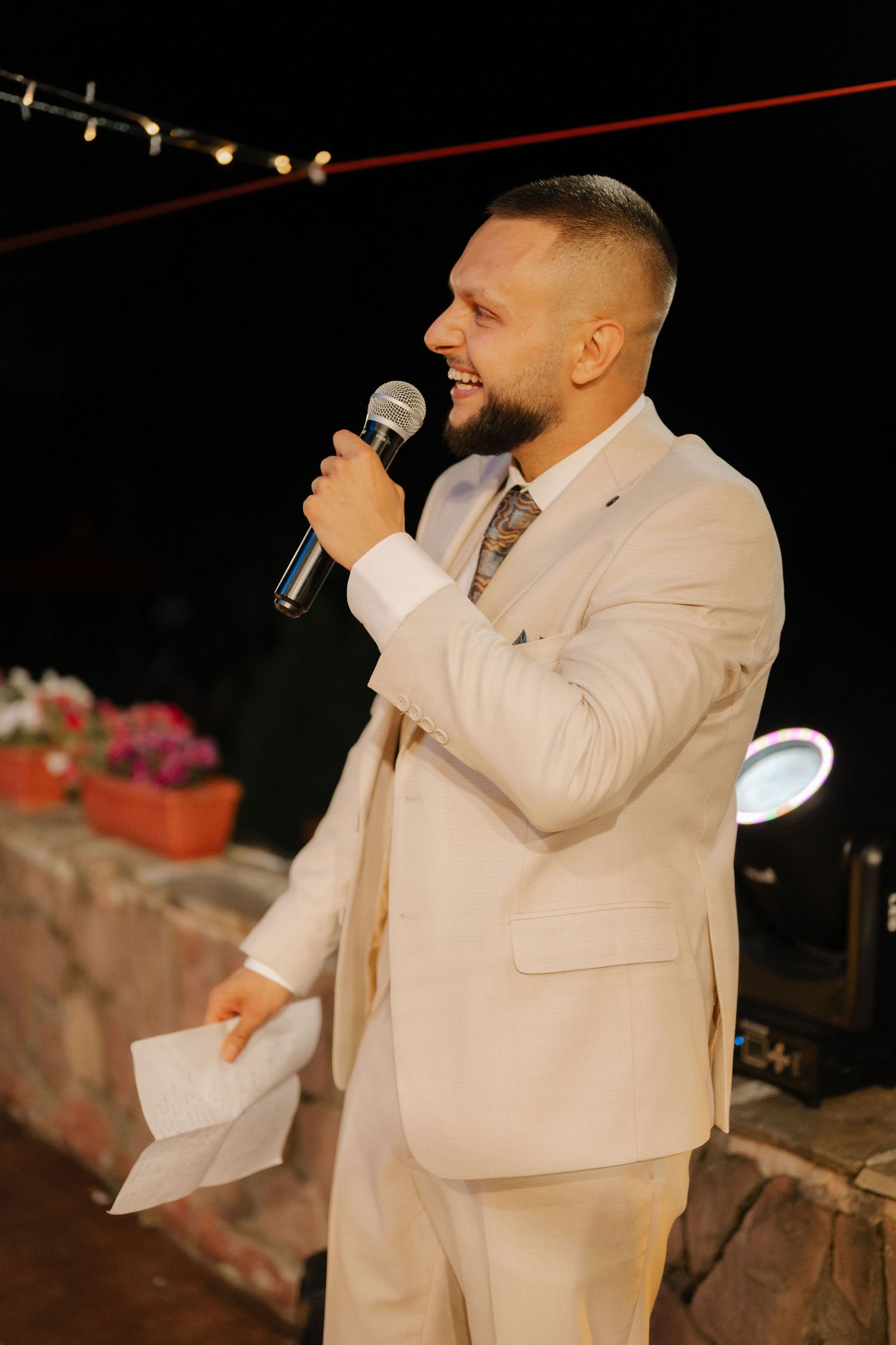 A person in a light beige suit gives a speech with a microphone at an outdoor evening event.