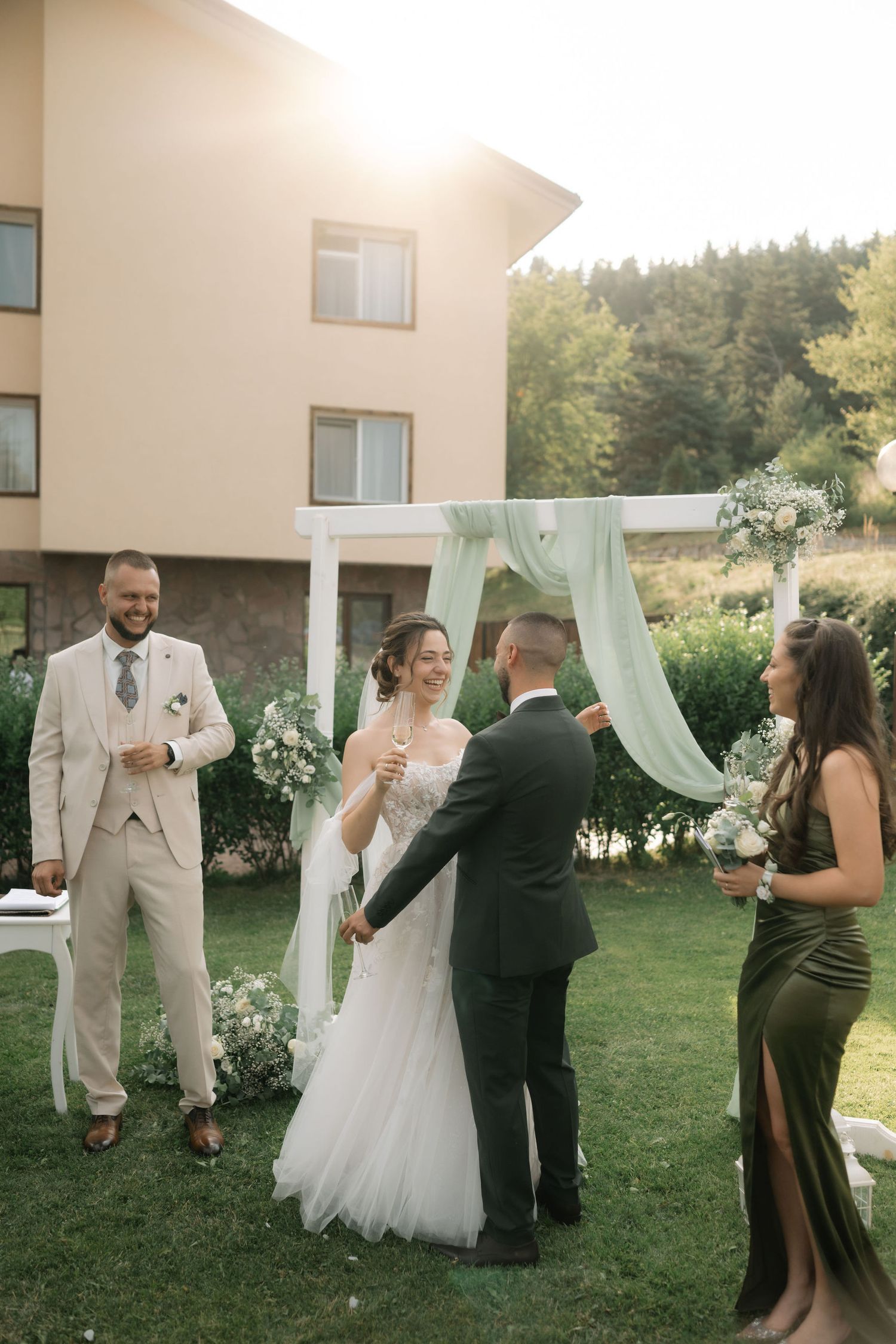 An outdoor wedding ceremony takes place on a lawn with flowing white drapery and floral decorations against a building backdrop.