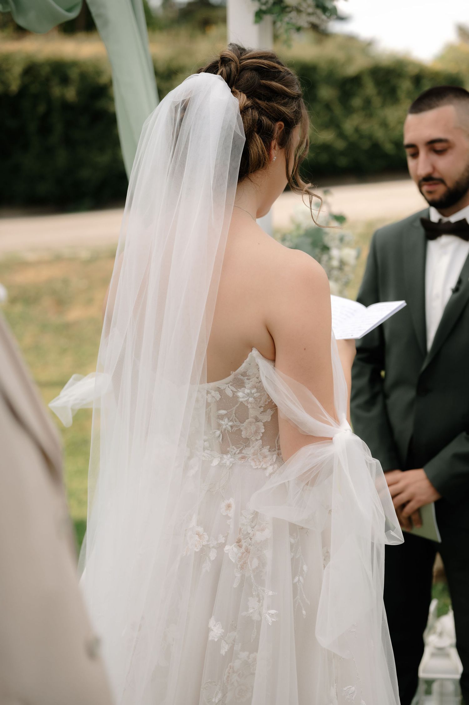 A wedding ceremony moment showing the back of a bride in a delicate lace gown with flowing veil during the vows.