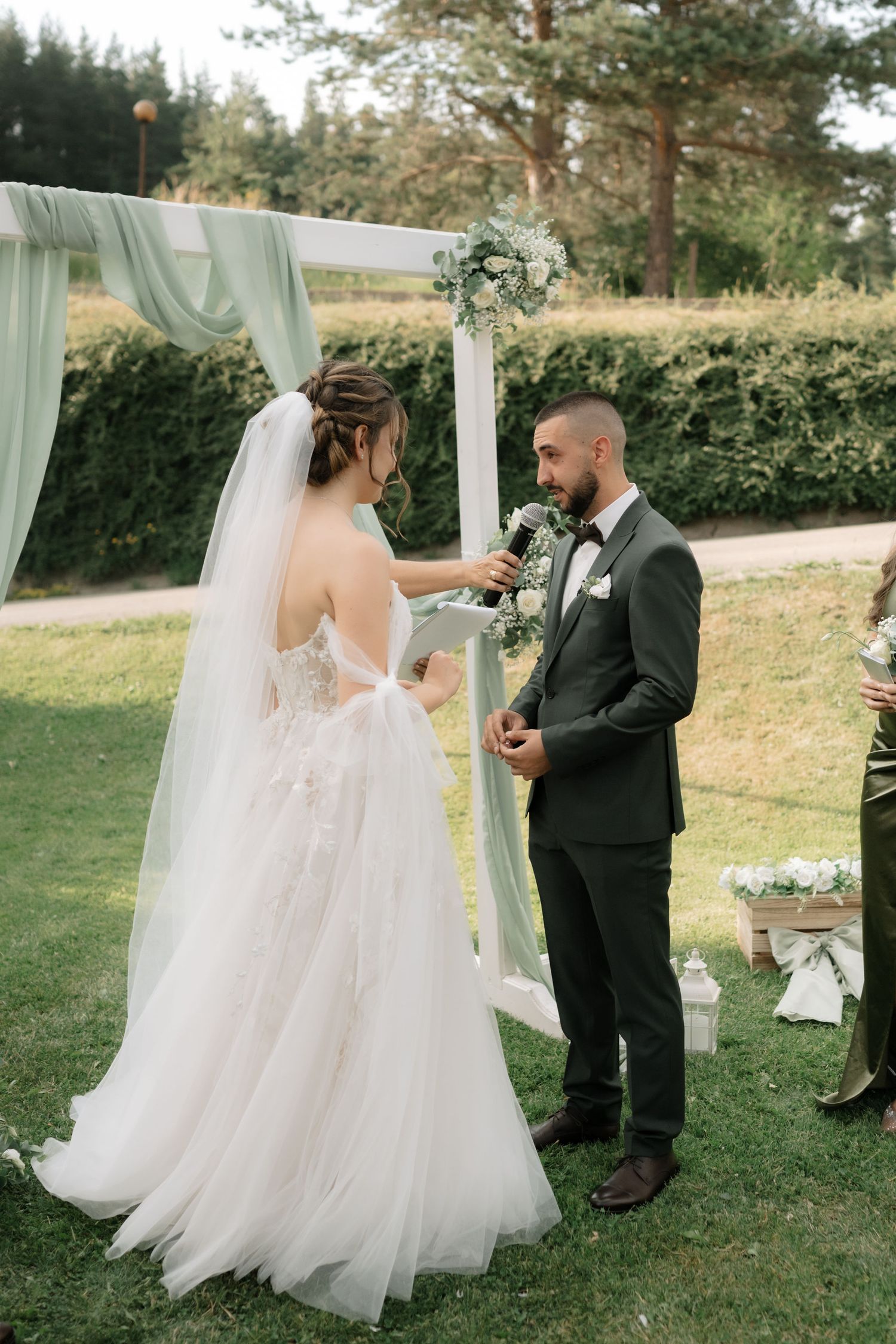 A romantic outdoor wedding ceremony takes place under a draped arch with floral arrangements in a garden setting.