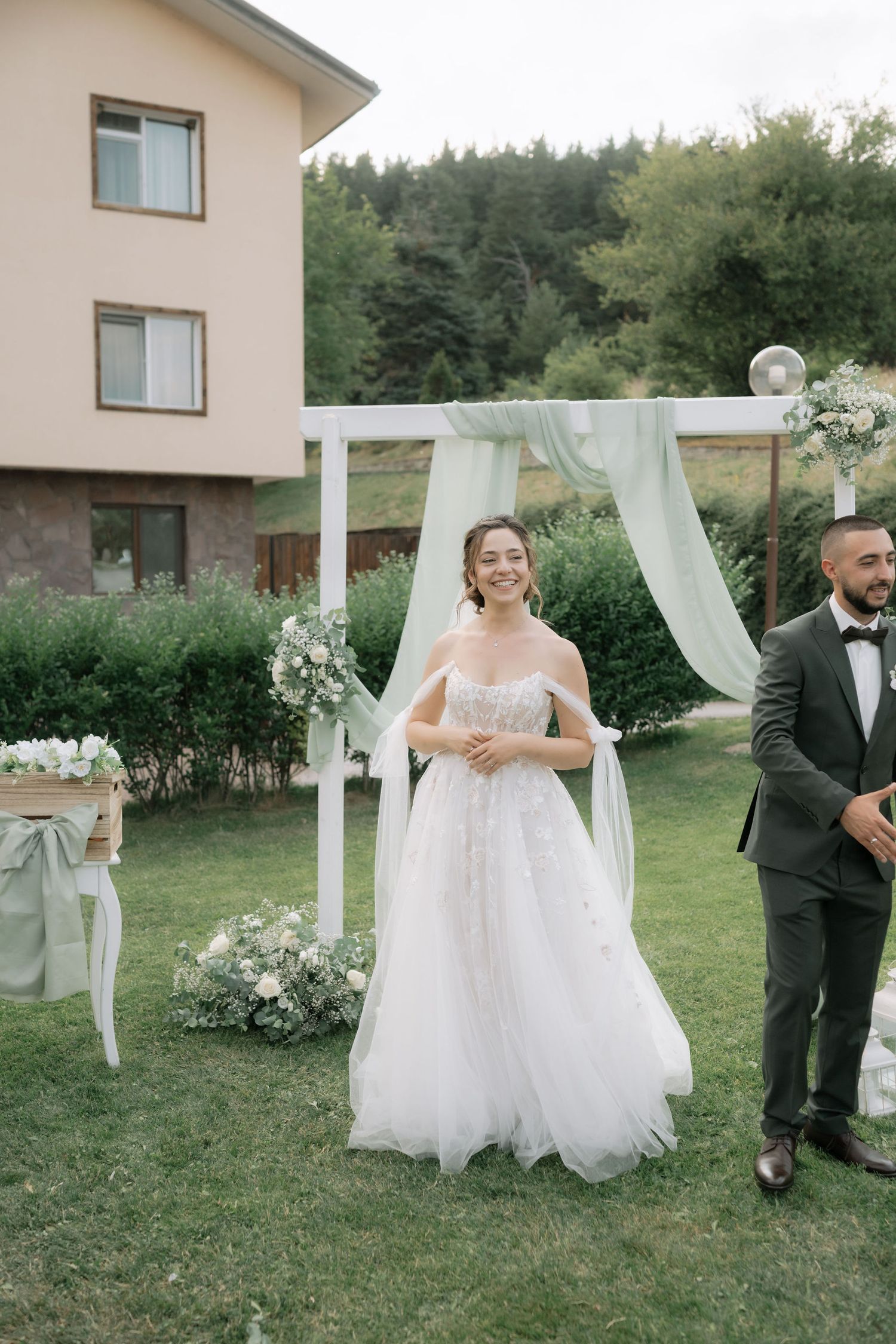 A bride and groom share a playful moment in a garden setting with white flowing fabric decor and greenery.