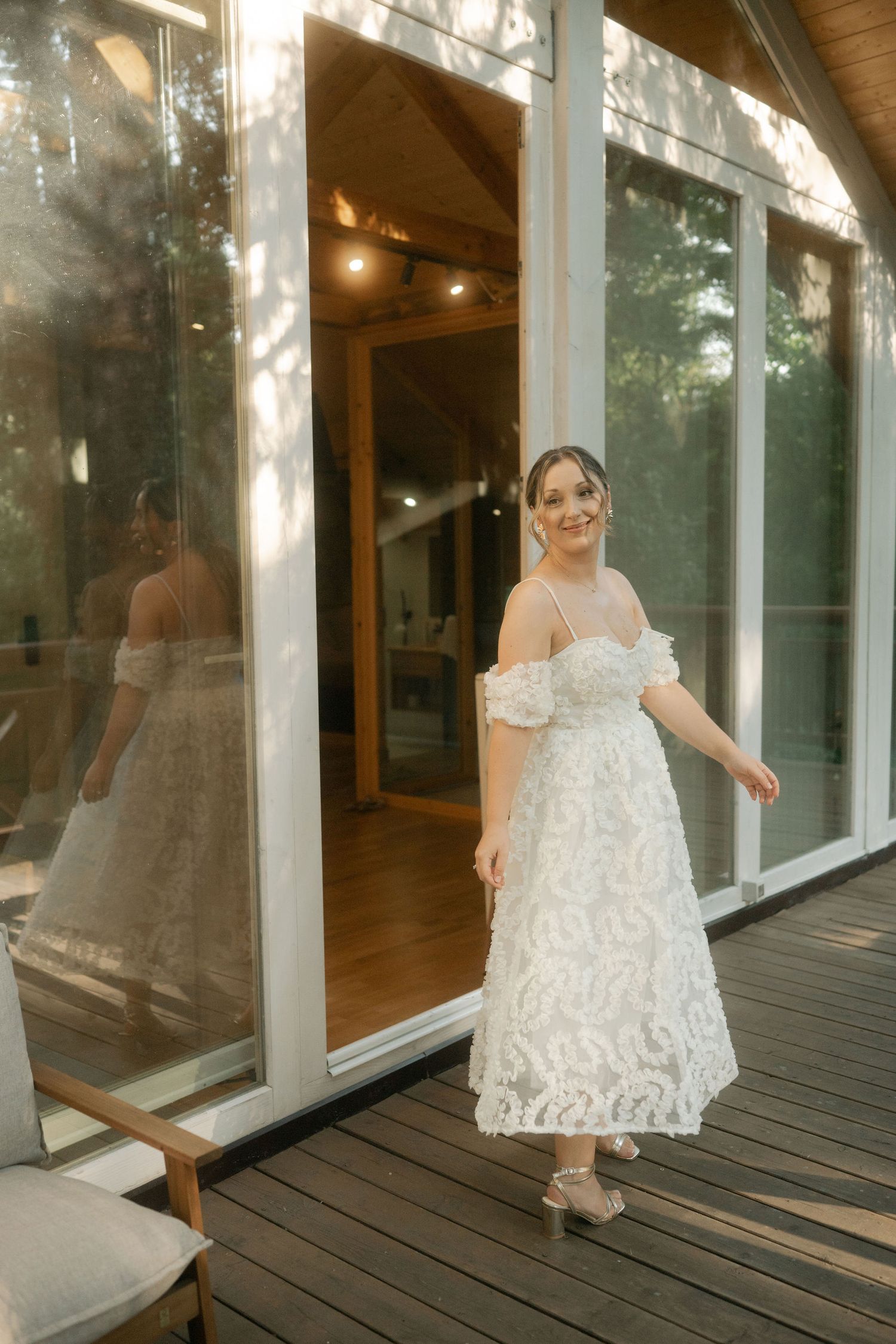 A white lace off-shoulder dress is worn on a wooden deck with glass windows reflecting the evening light.