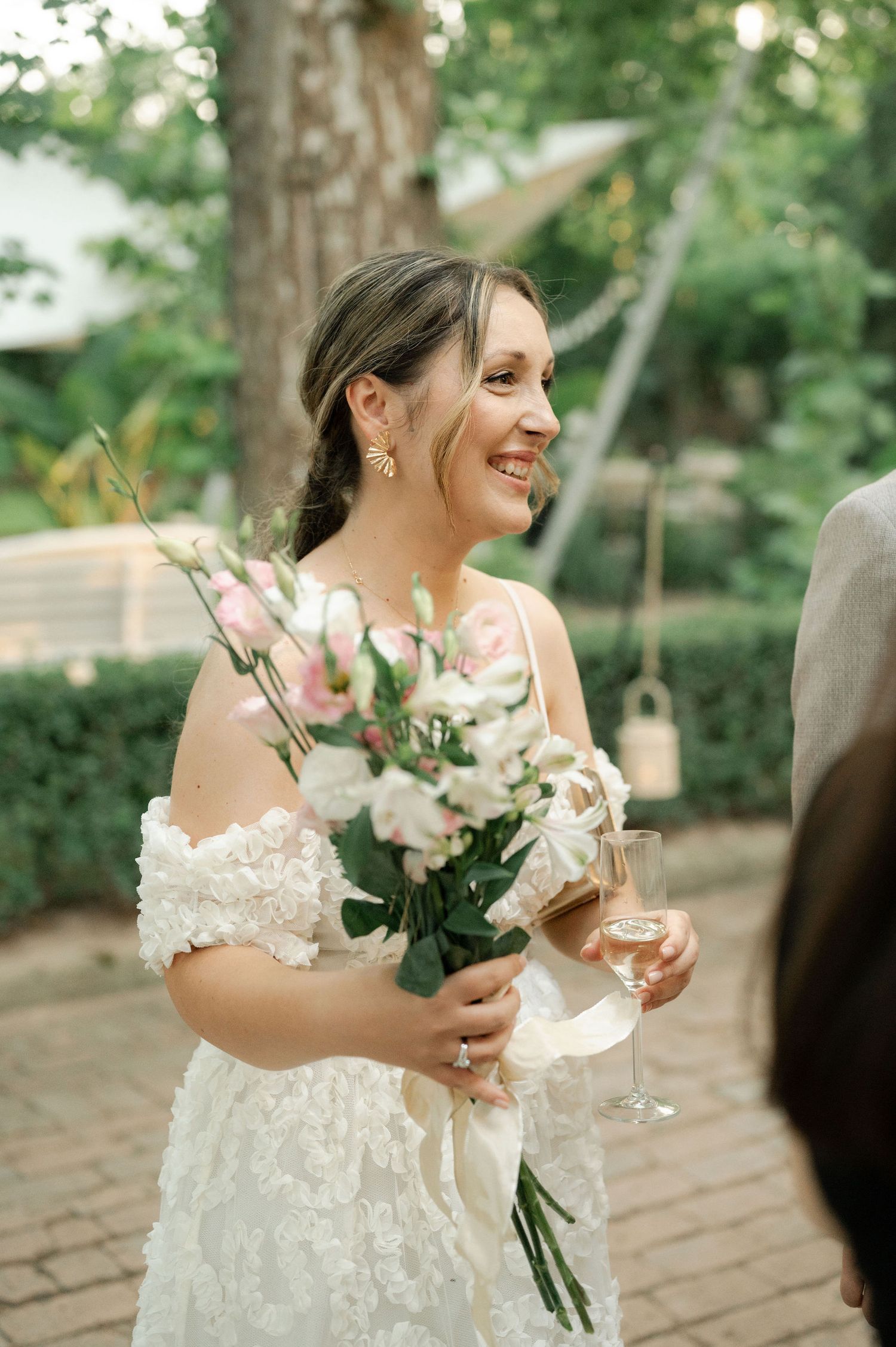A bride in a white lace dress smiles while holding a bouquet at an outdoor wedding ceremony.