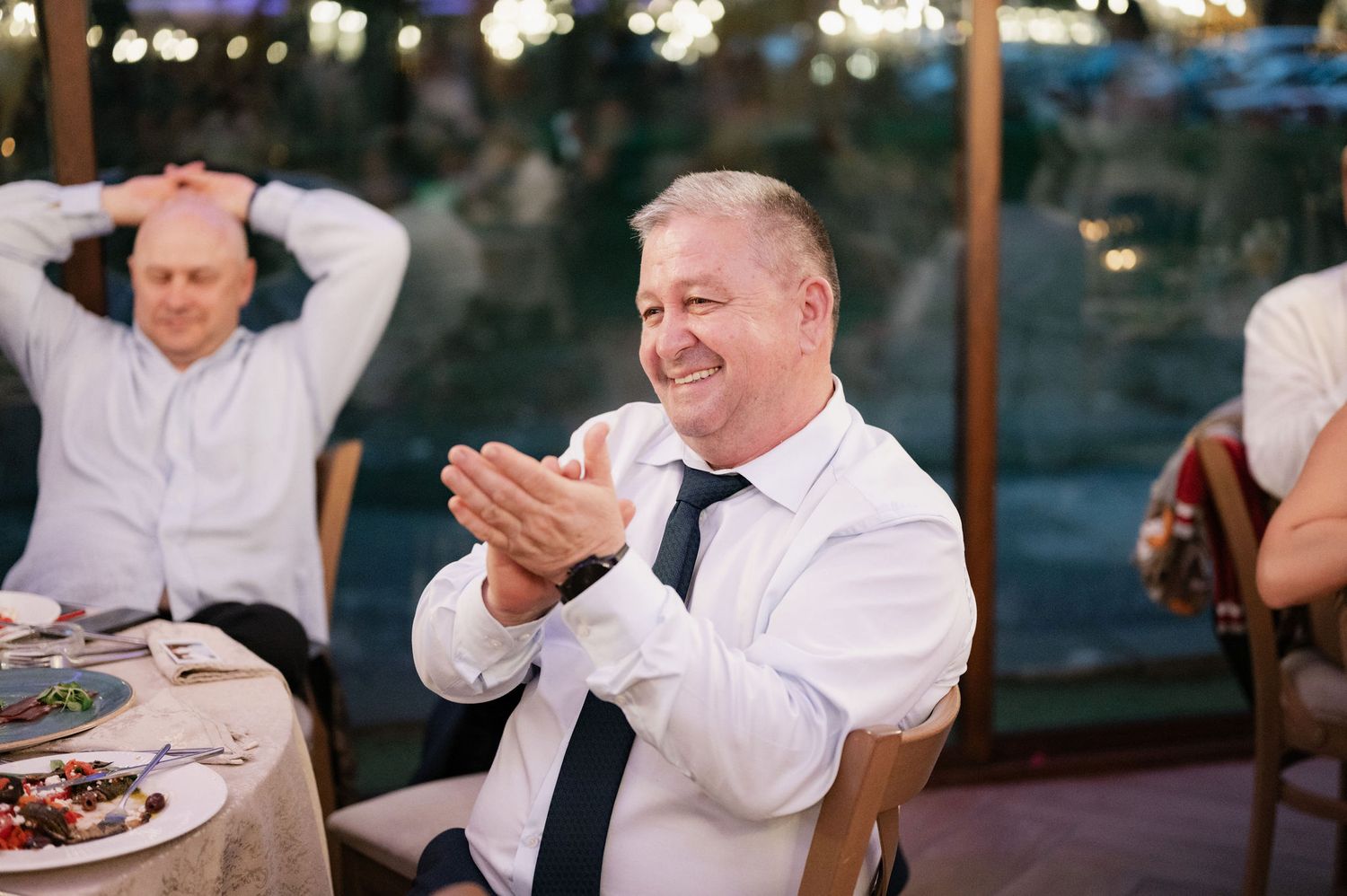 People in formal attire laugh and socialize during an outdoor evening celebration with twinkling lights in background.