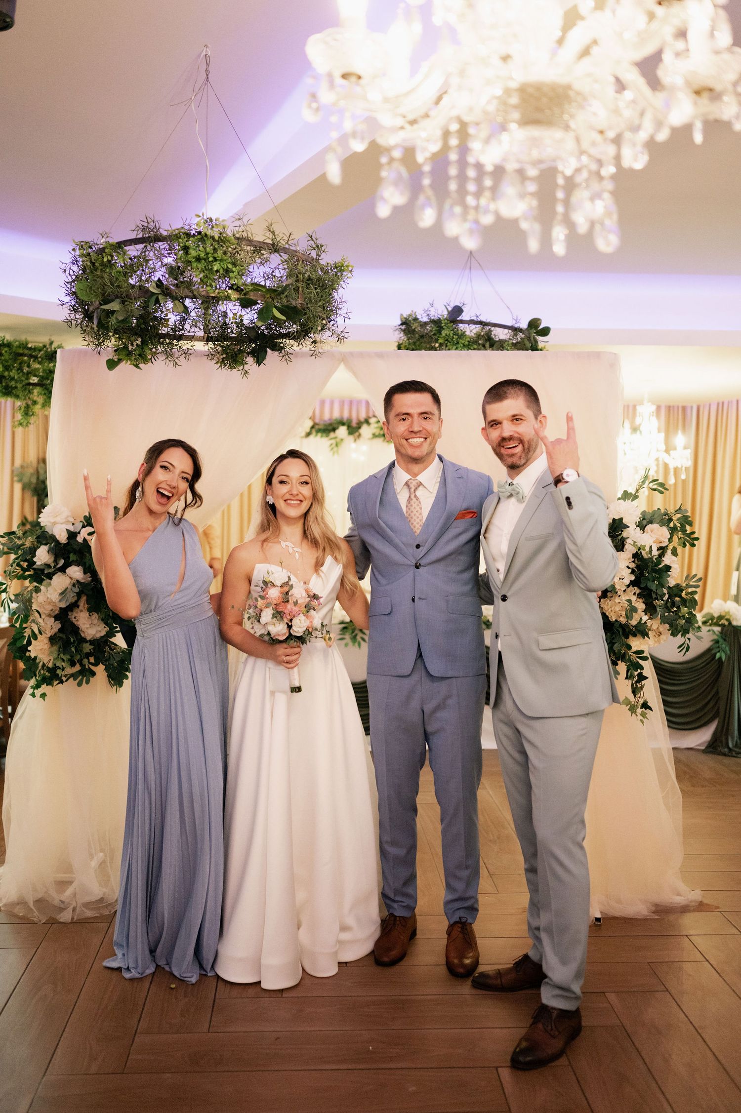 Wedding party in gray and white attire poses under elegant chandeliers in a reception venue.