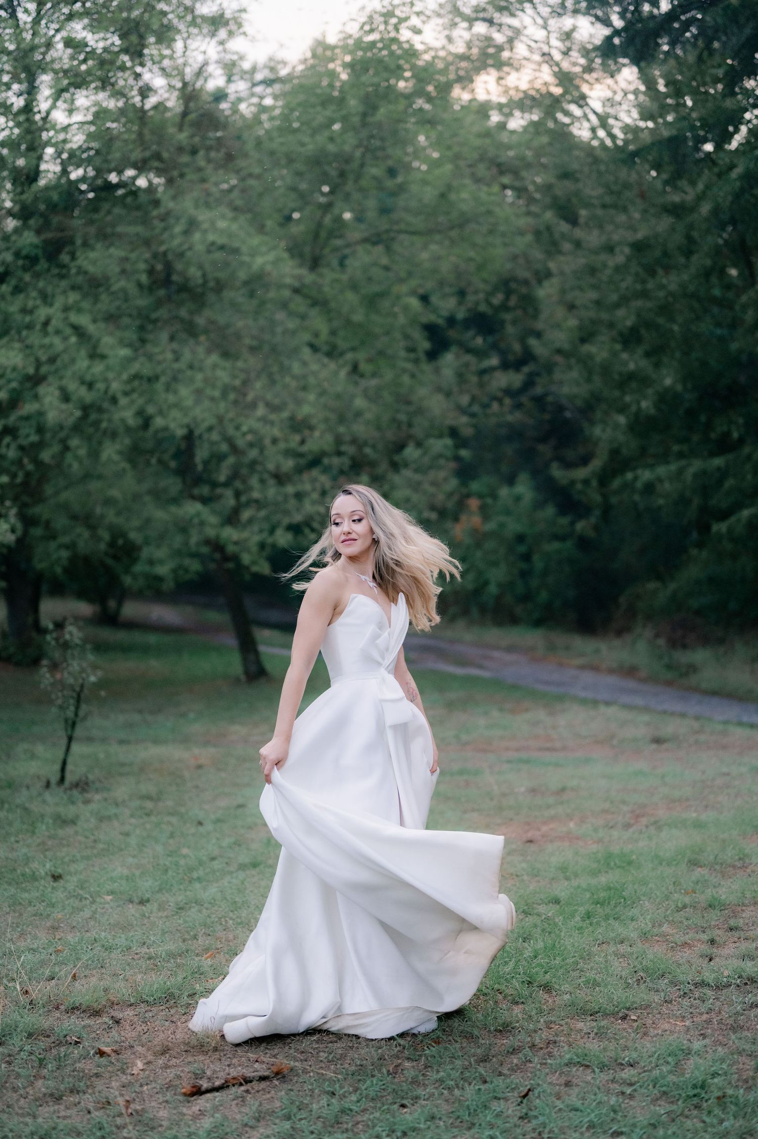 A bride in a flowing white wedding gown twirls gracefully in a serene outdoor setting surrounded by trees at dusk.