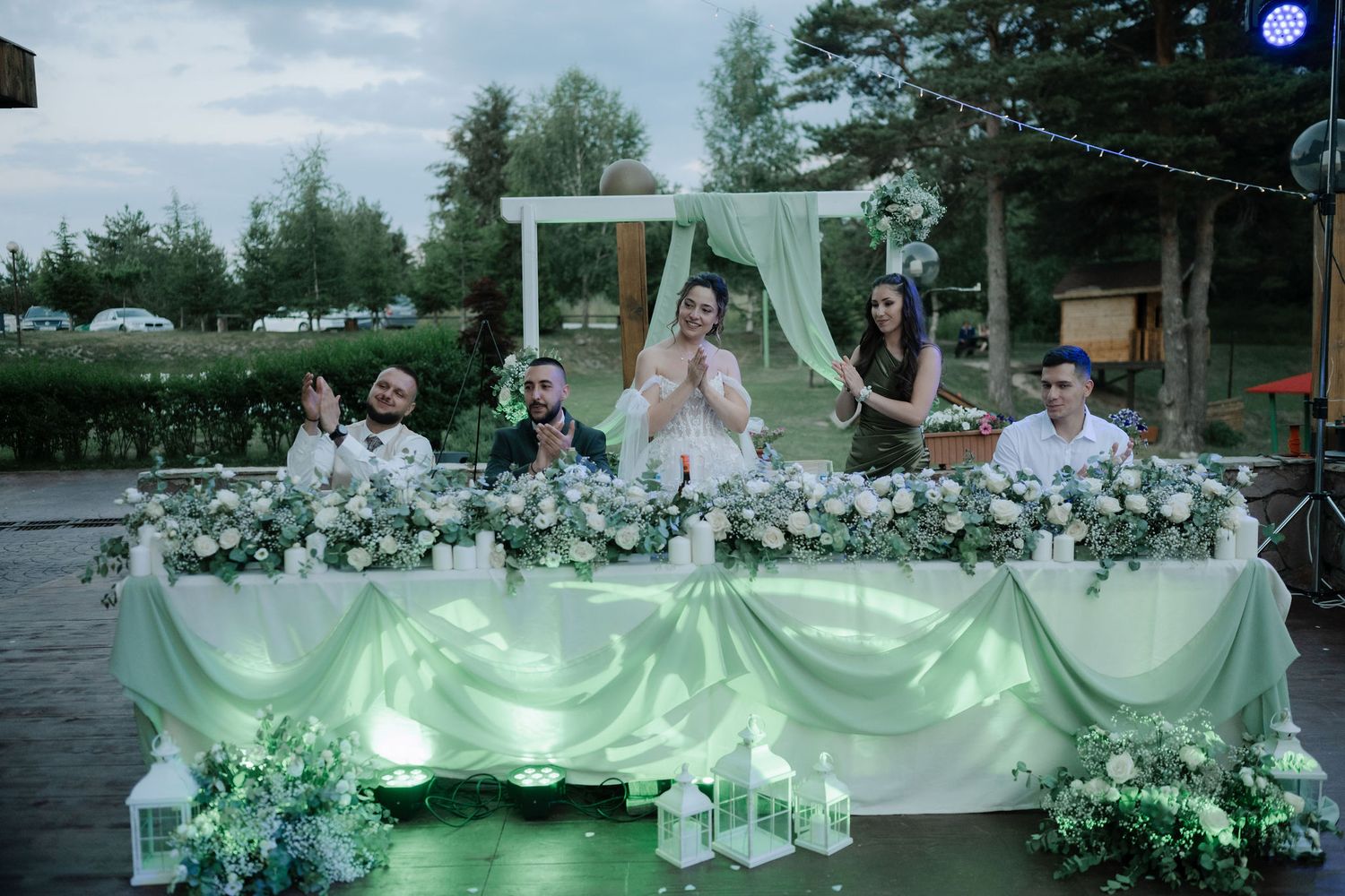 Wedding party seated at an elegant white table decorated with lush greenery and glowing candles at an outdoor reception.