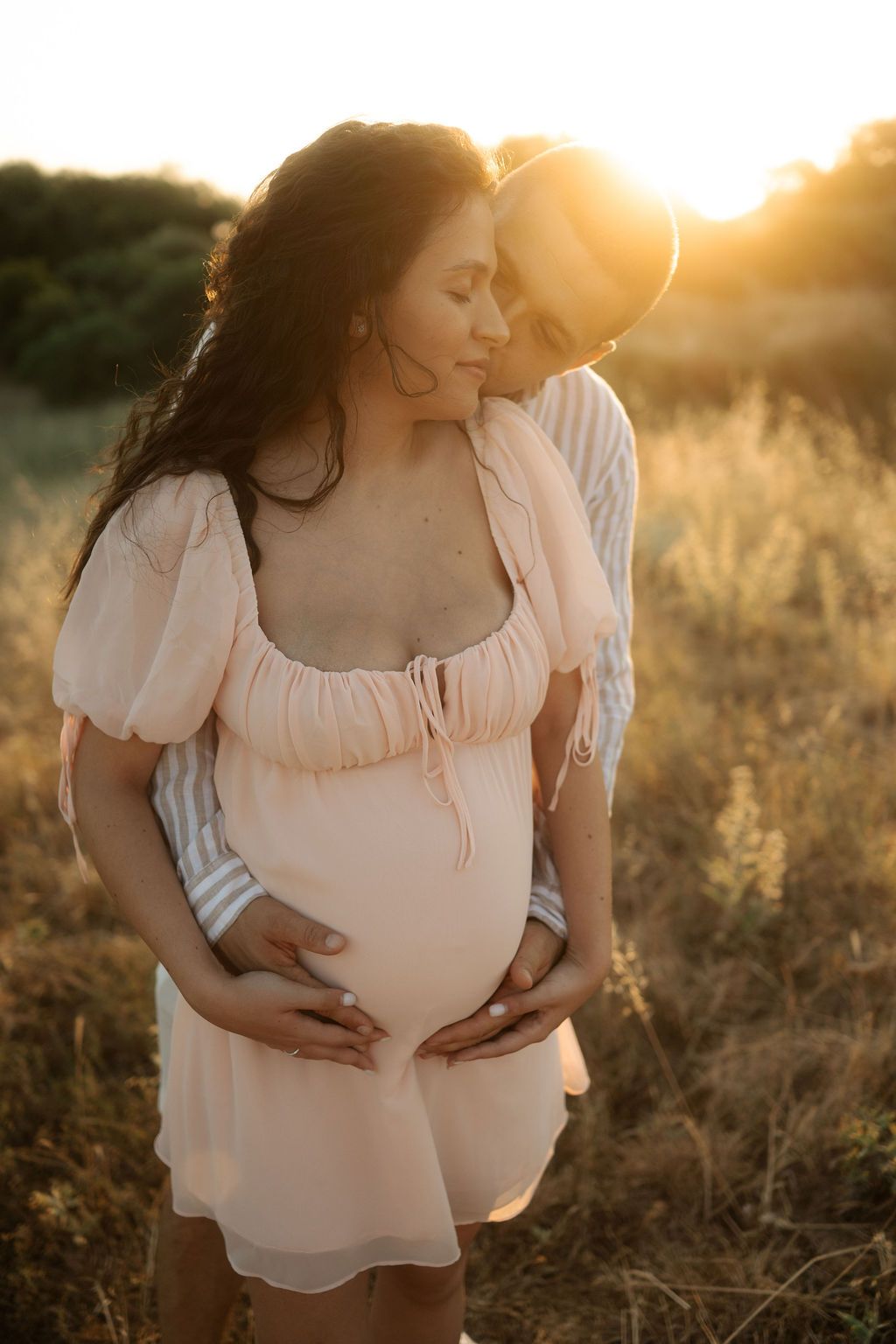 Couple shares intimate moment in warm sunset lighting while standing in field of dried grass.
