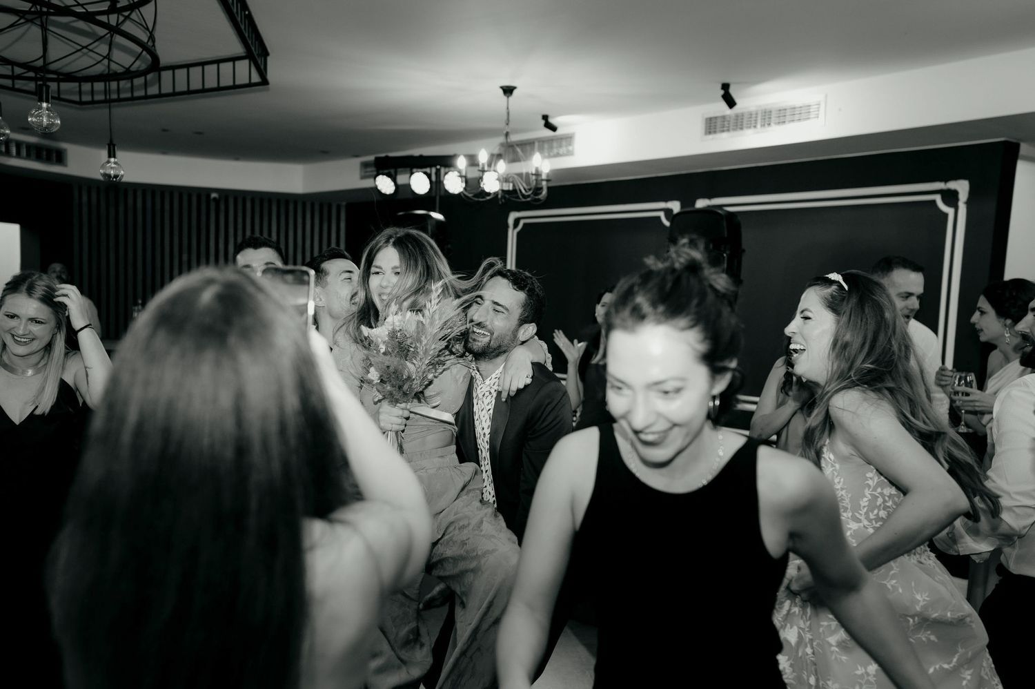 A group of people dance and celebrate together in what appears to be a dimly lit party or nightclub setting.